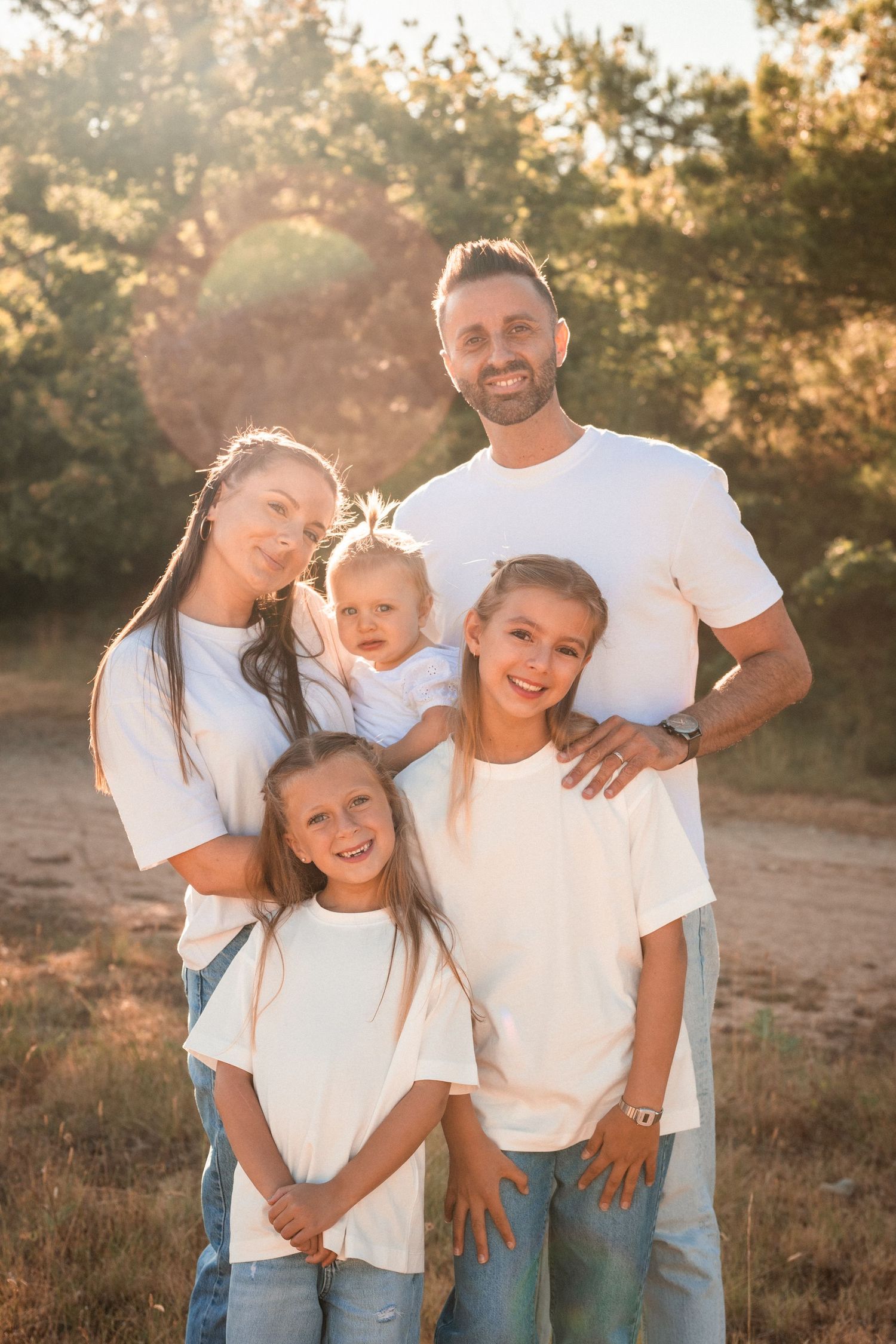 Happy family of five in matching white shirts posing together outdoors in golden sunlight.