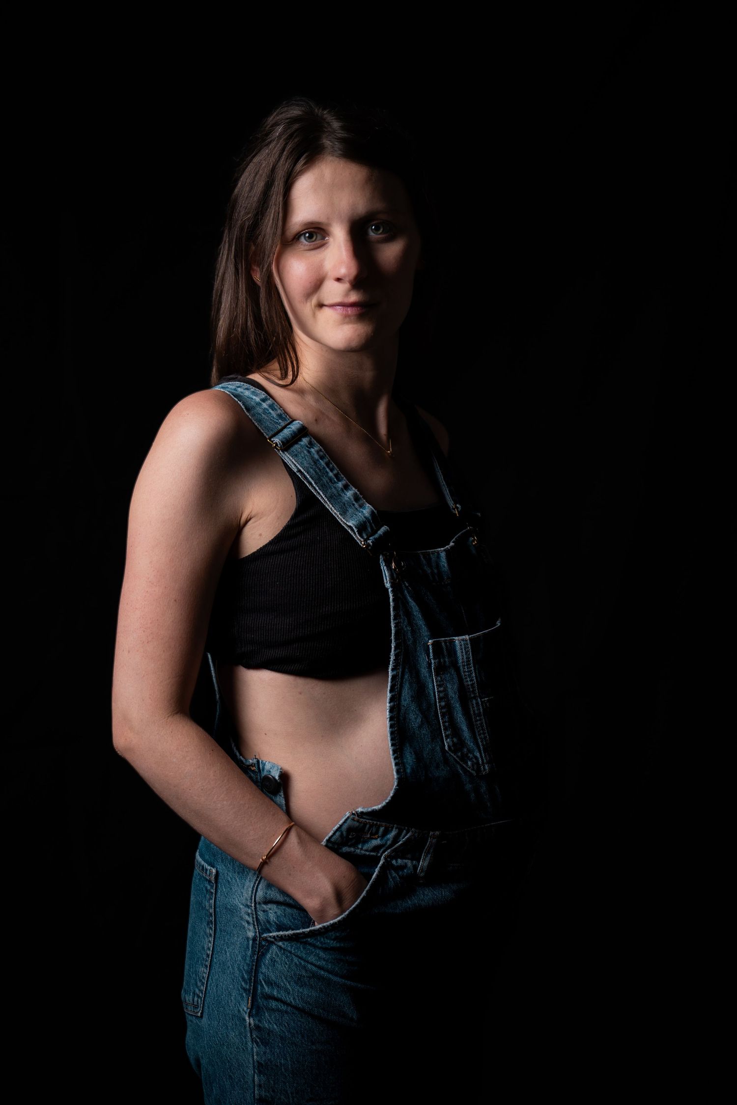 Pregnant woman in denim overalls and black crop top posing confidently against a dark black studio background.
