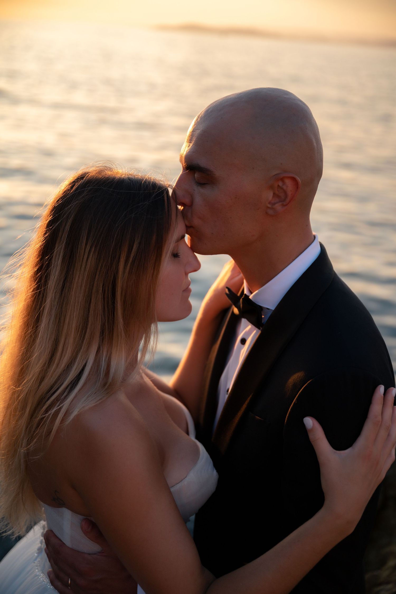 Groom in tuxedo kisses bride on forehead during golden sunset by the sea.