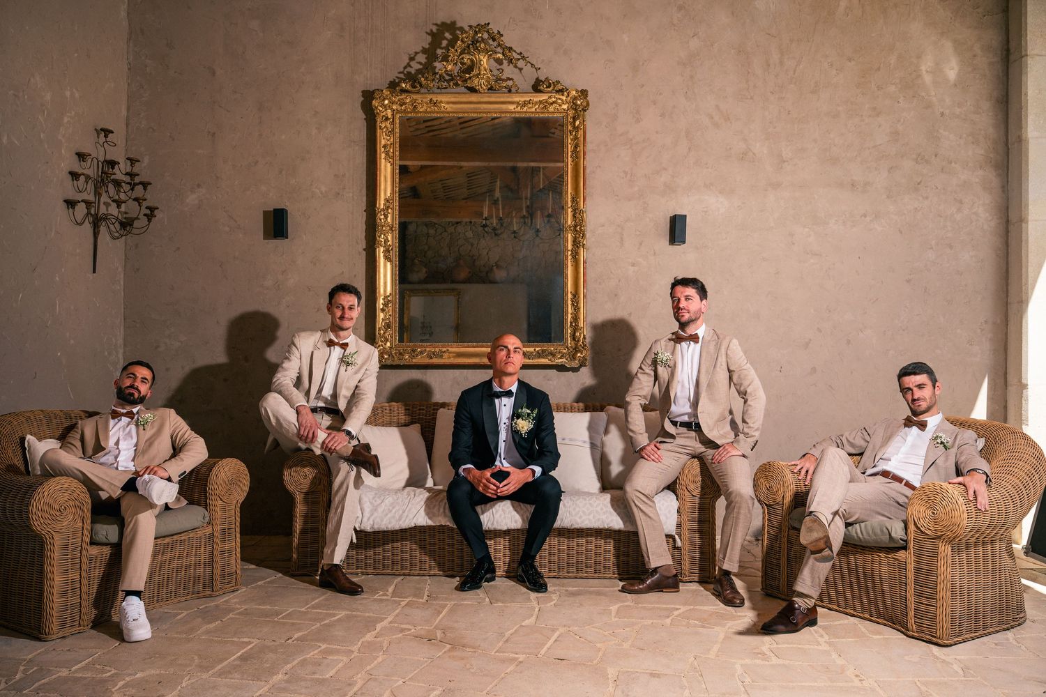 Groom and groomsmen in tan suits pose elegantly in a rustic venue with wicker chairs and ornate gold mirror.