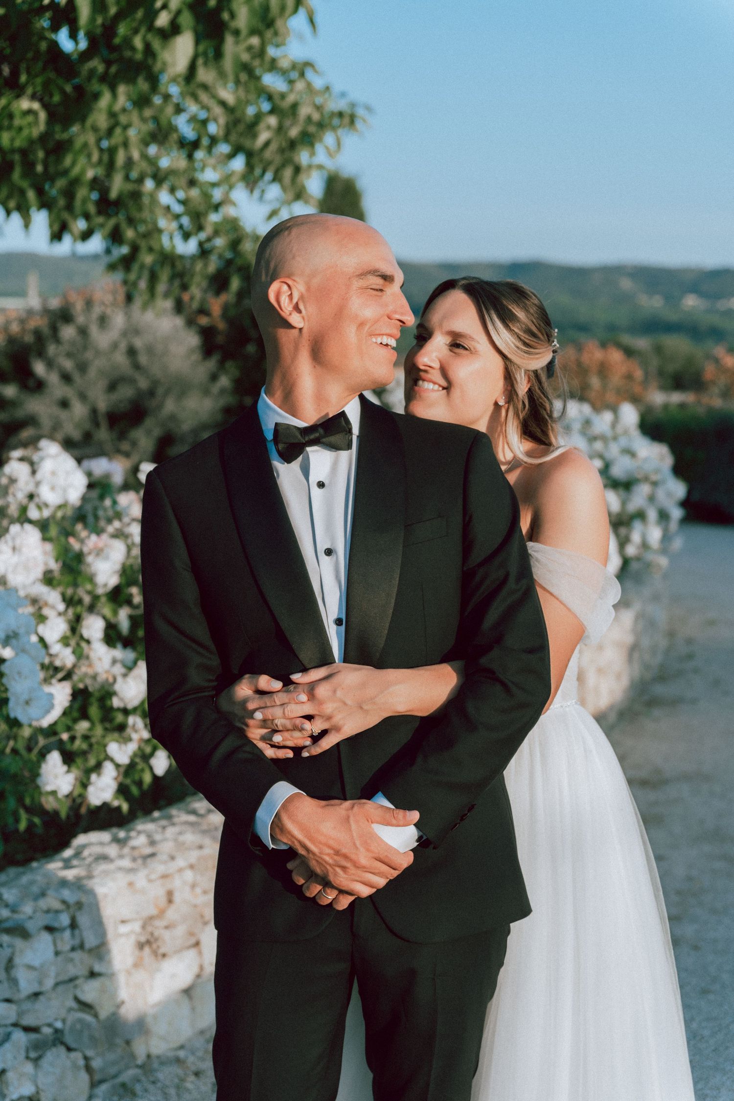 Bride embraces groom from behind as they smile together in garden wedding portrait.