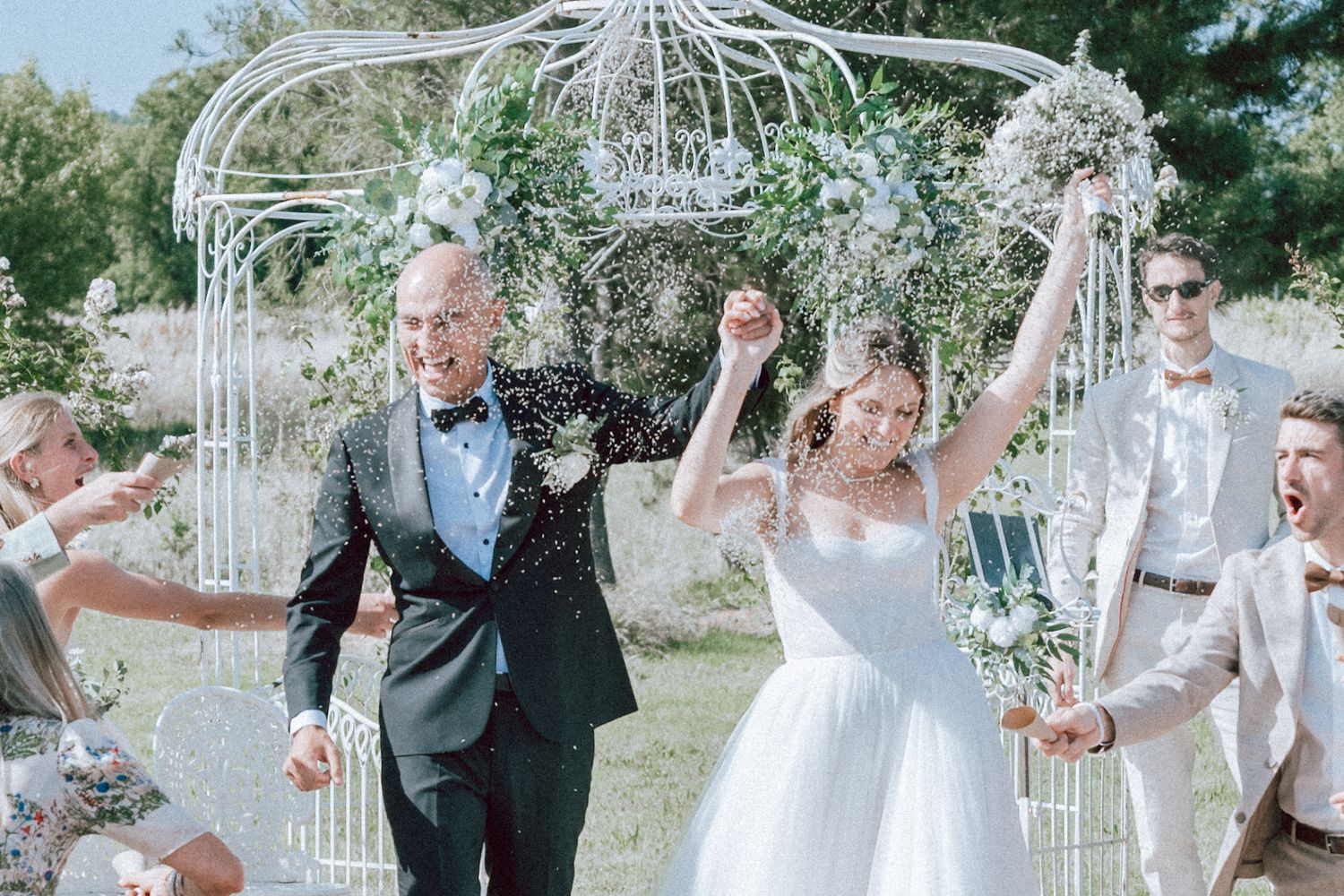 Joyful newlyweds celebrate with confetti under a white garden gazebo surrounded by cheering guests.