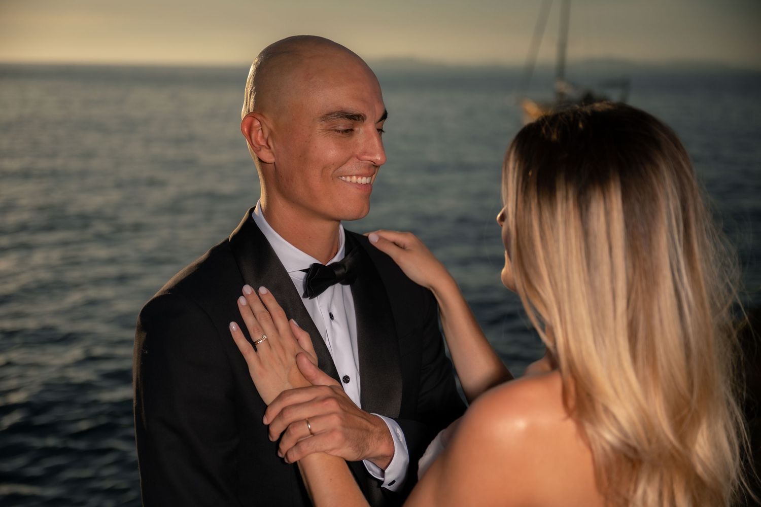 Couple in formal wear sharing a romantic moment by the ocean at sunset, man in tuxedo smiling at blonde woman.