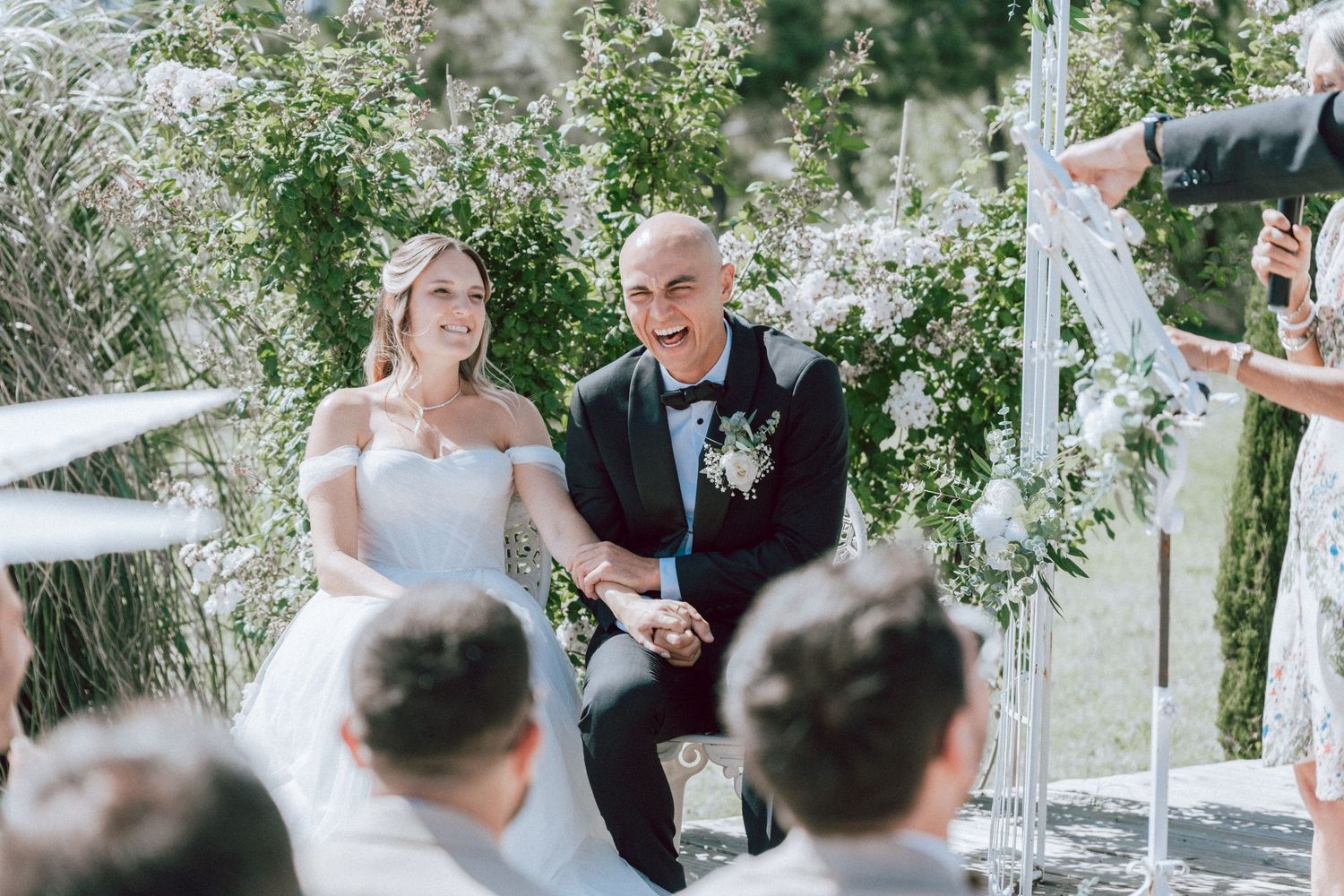 Smiling bride and groom kneeling together during outdoor wedding ceremony surrounded by greenery.
