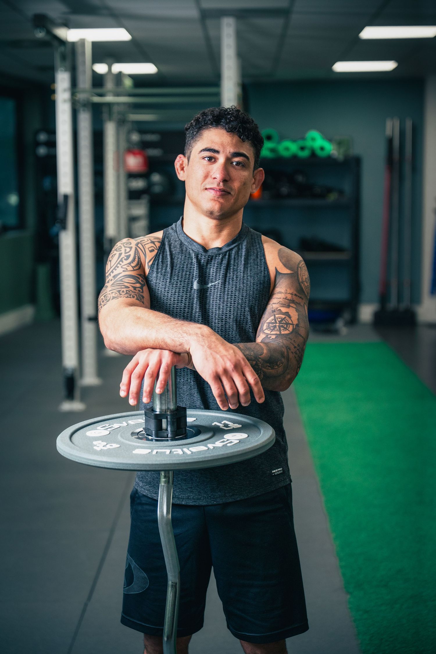 Tattooed athletic man leaning on a barbell weight plate in a modern gym.
