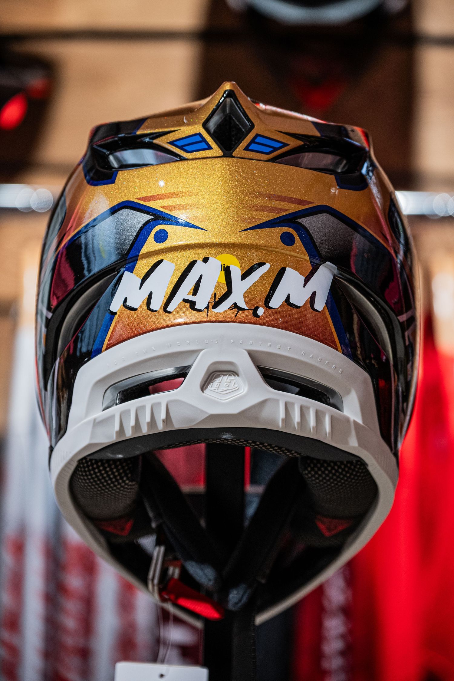Gold and black MAX.M motocross helmet with blue accents displayed on a stand in a motorcycle gear shop.