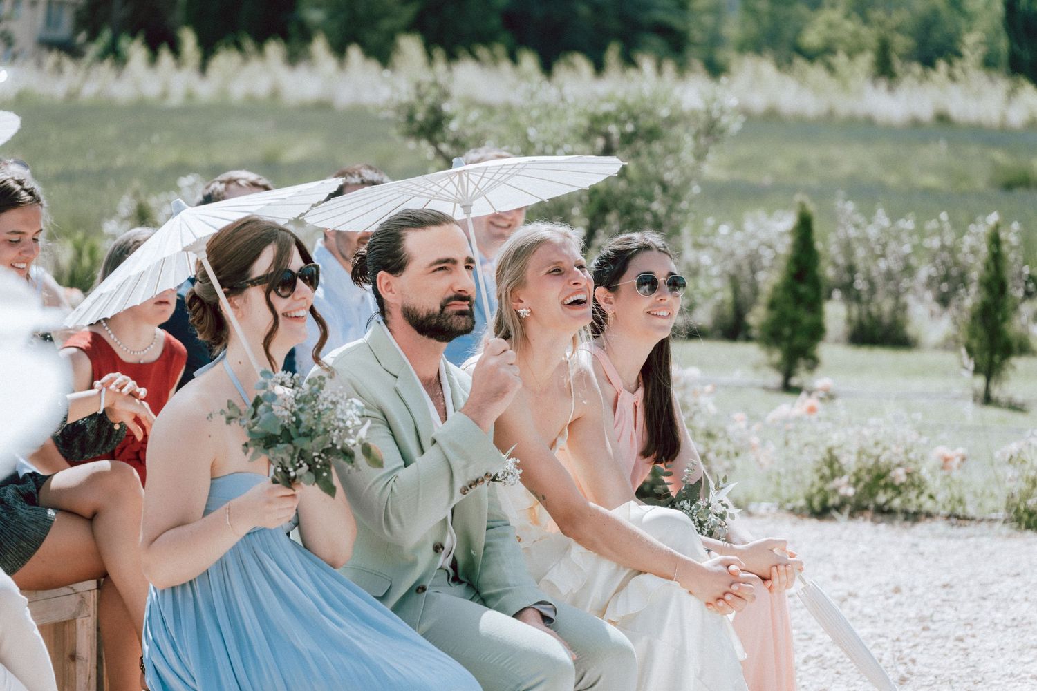 Wedding guests laugh and smile while seated outdoors holding white parasols during sunny ceremony.