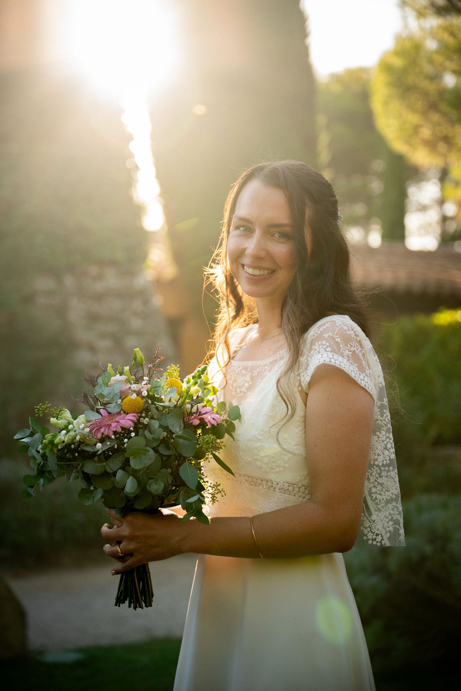 Smiling bride in lace wedding dress holding colorful bouquet with golden sunlight glowing behind her outdoors.