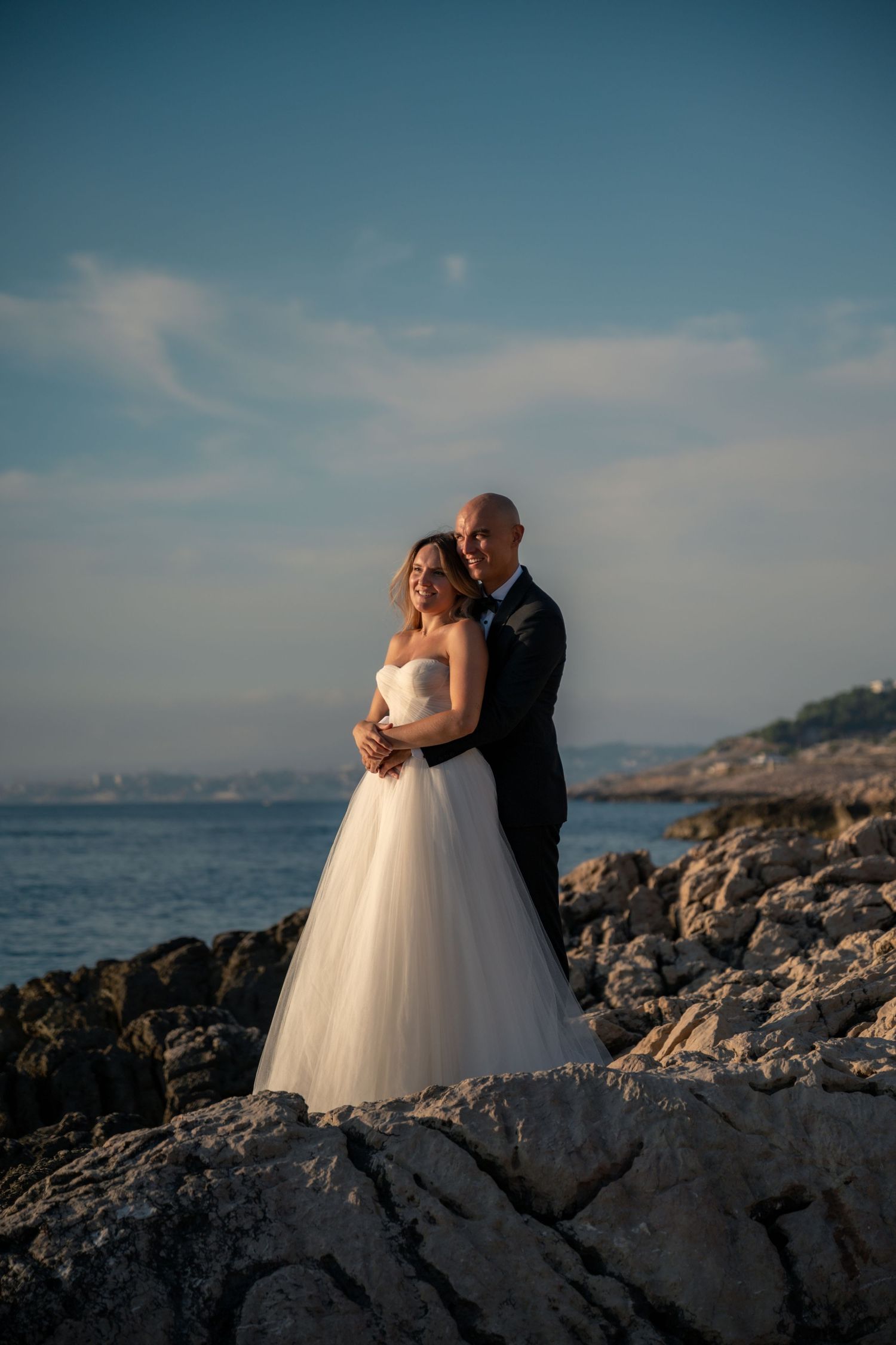 Bride and groom embracing on rocky seaside cliffs at golden hour sunset.