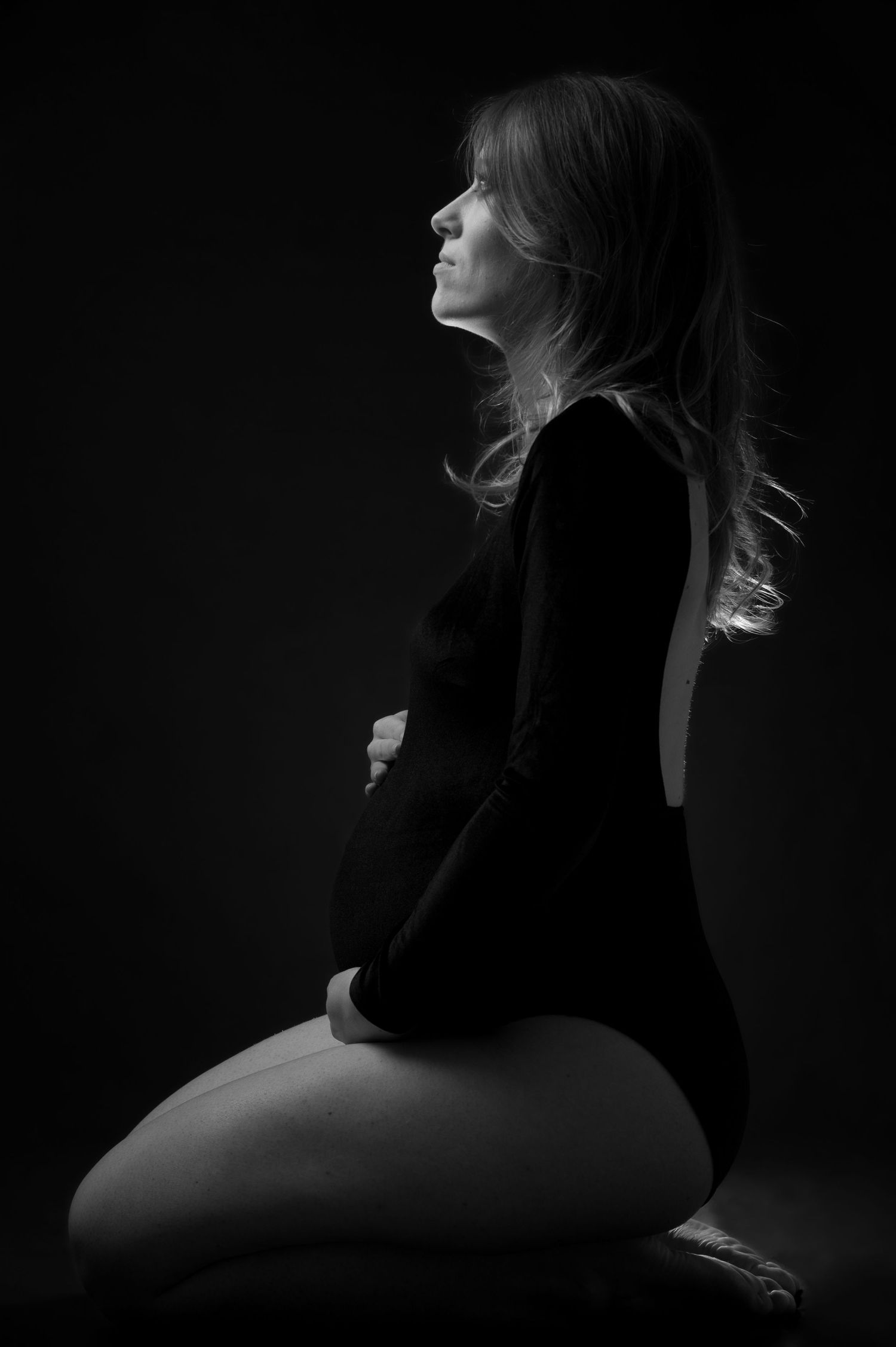 Black and white profile portrait of a pregnant woman kneeling, cradling her baby bump in a dark studio setting.