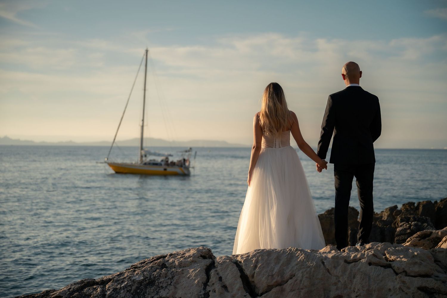 Bride and groom hold hands on rocky shore watching a sailboat at sunset.