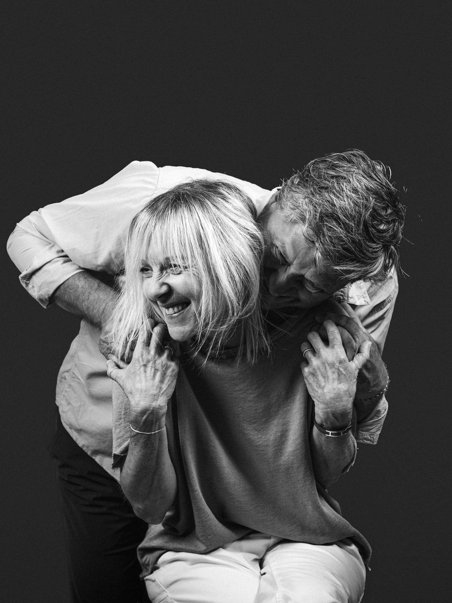 Black and white portrait of an older couple laughing playfully together in a studio against a dark background.