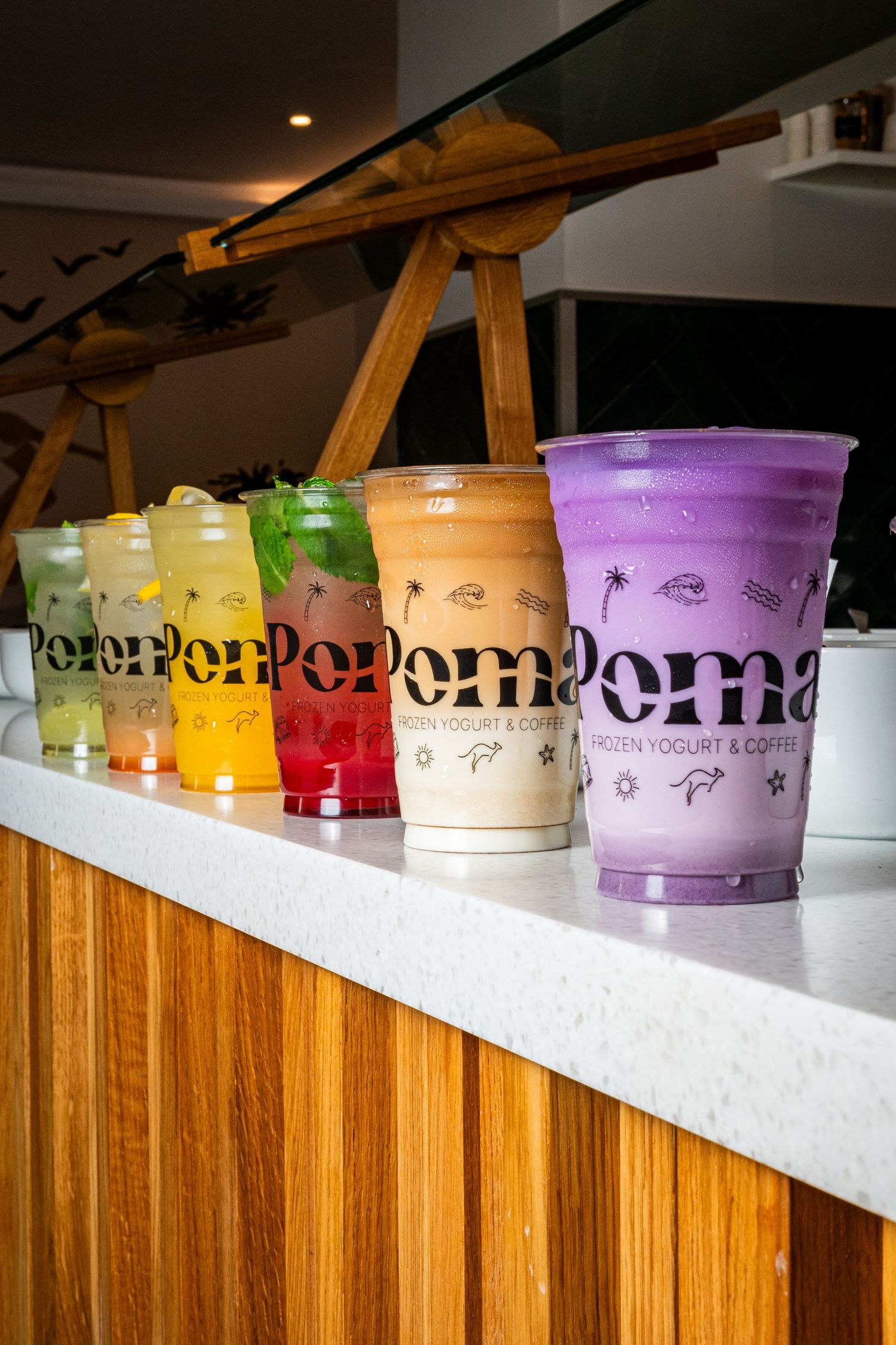 Row of colorful Poma Frozen Yogurt & Coffee drinks displayed on wooden counter.