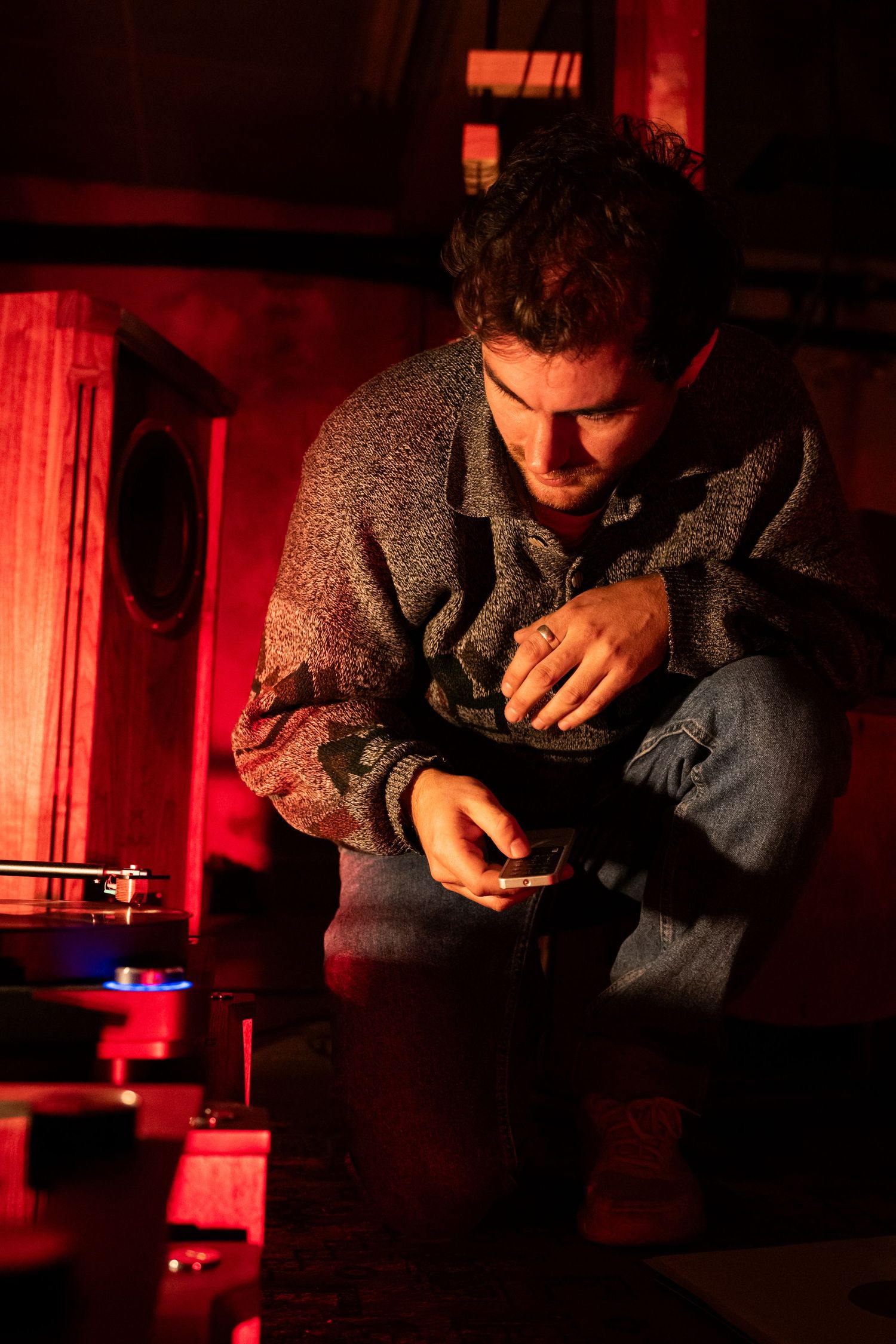 Man in sweater and jeans sits in red-lit room near a turntable, focused on handling a vinyl record.
