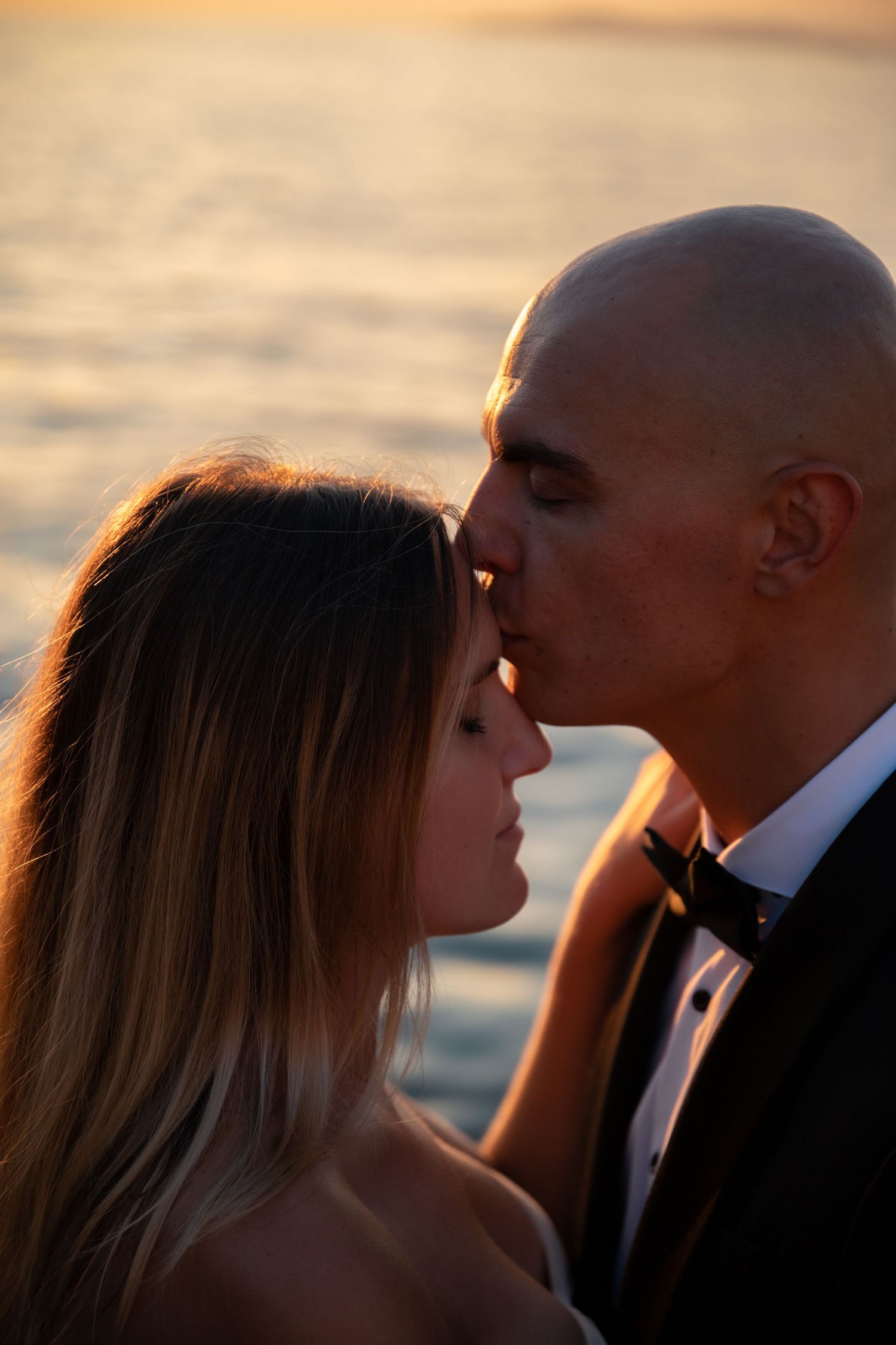 Groom in tuxedo kissing bride on forehead at golden sunset by the sea.