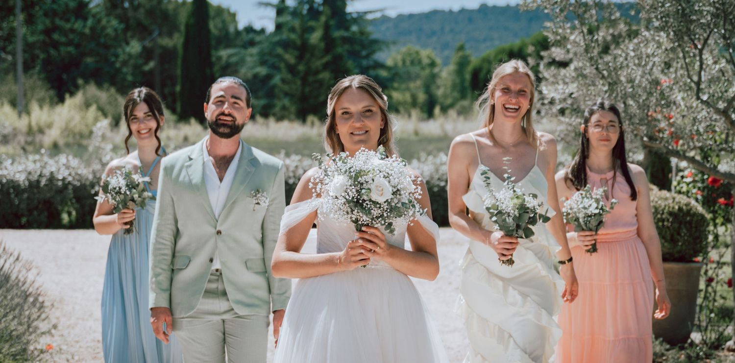 Bride and groom with bridesmaids holding white bouquets outdoors at a sunny wedding ceremony.