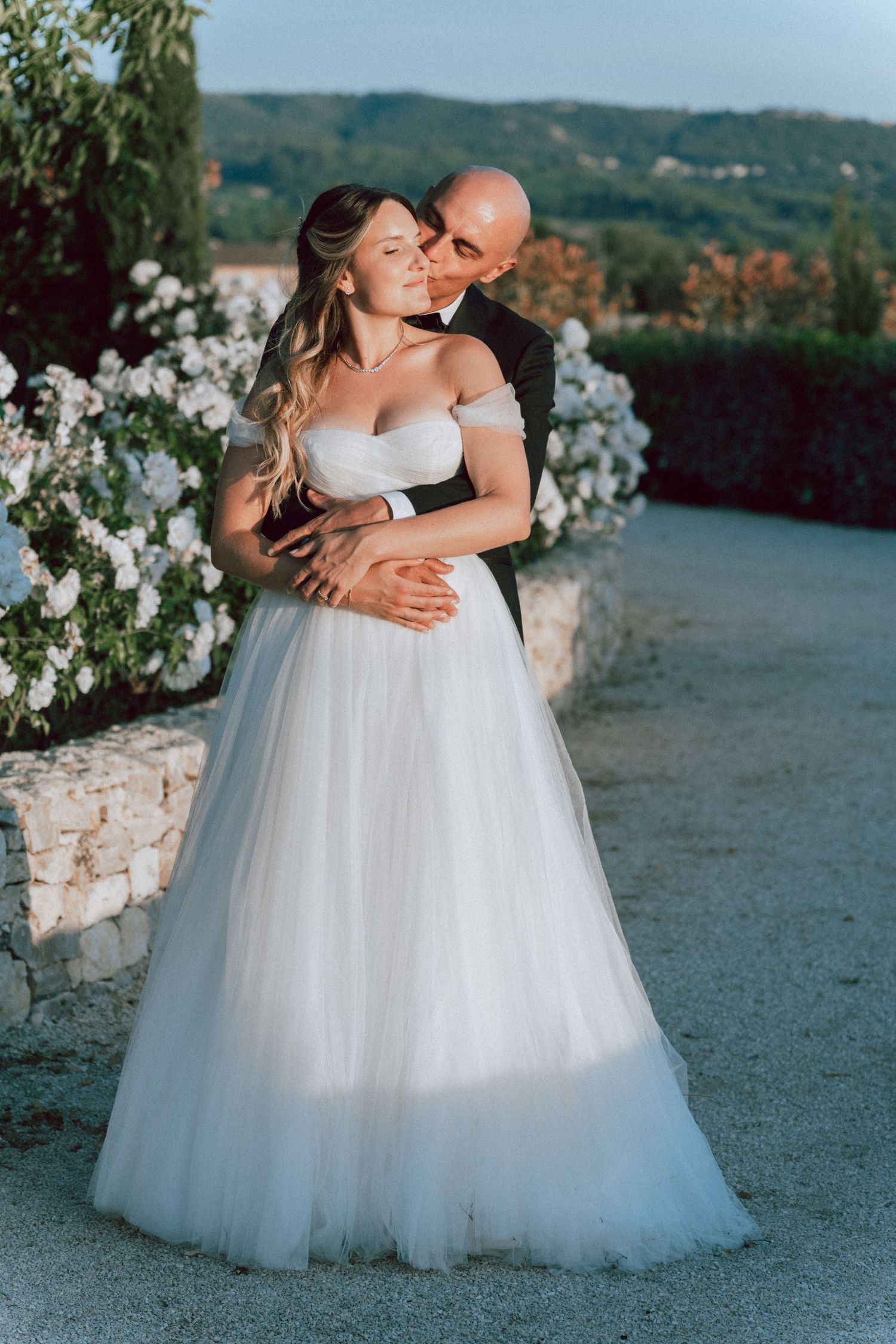 Groom embracing bride from behind near blooming flowers, bride in elegant off-shoulder tulle wedding dress.