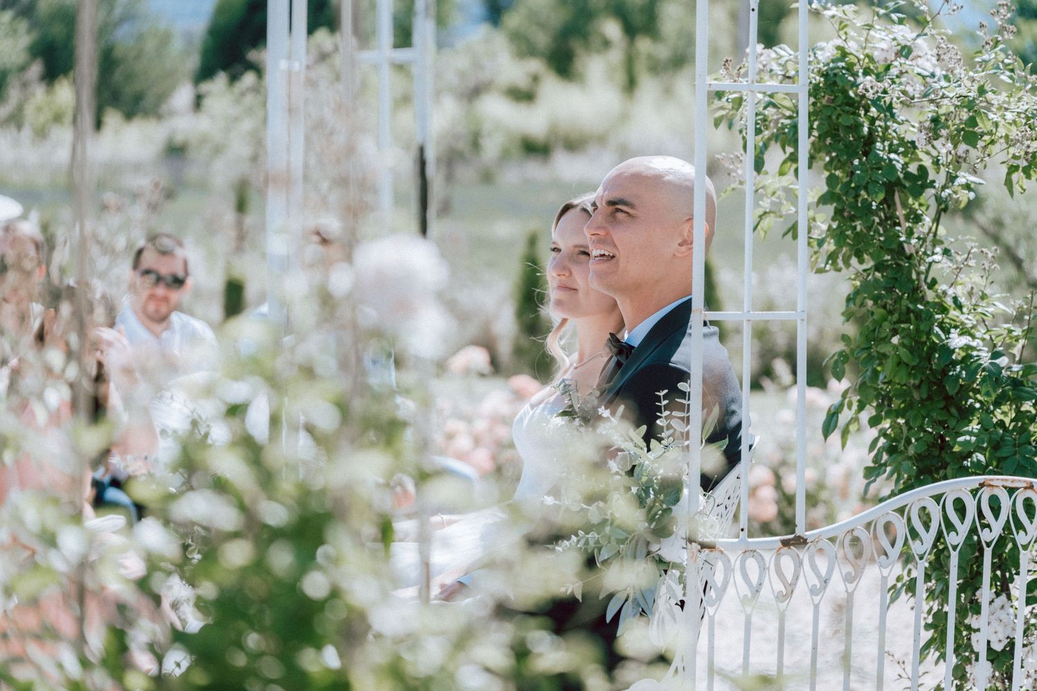 Wedding guests seated and watching outdoor ceremony through decorative white iron gazebo with flowers.
