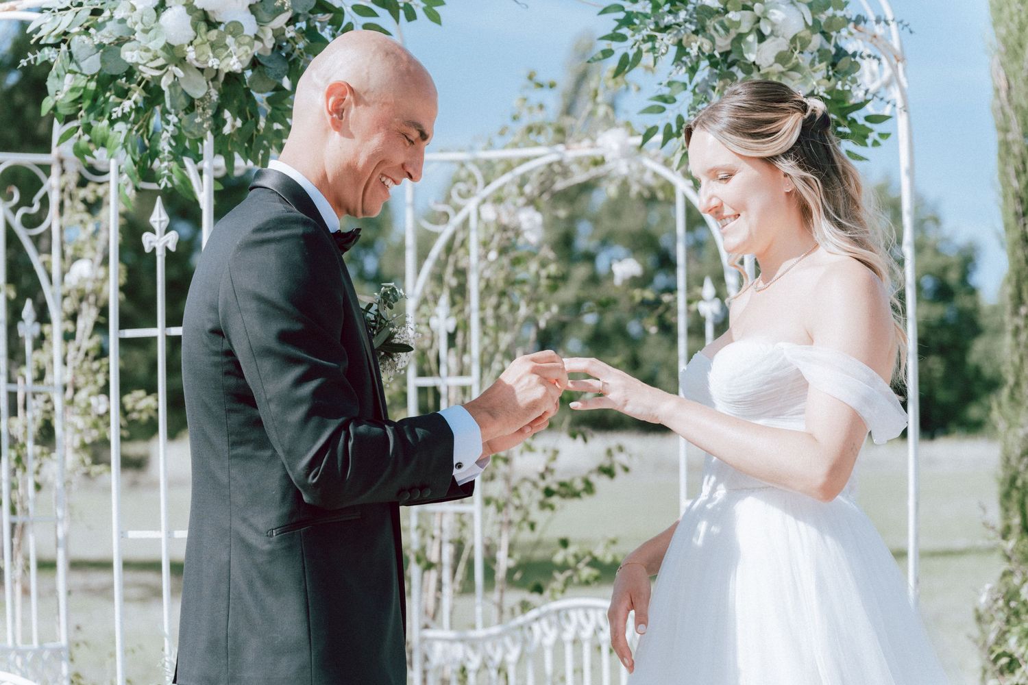 Groom places wedding ring on brides finger at outdoor ceremony beneath white floral arch.