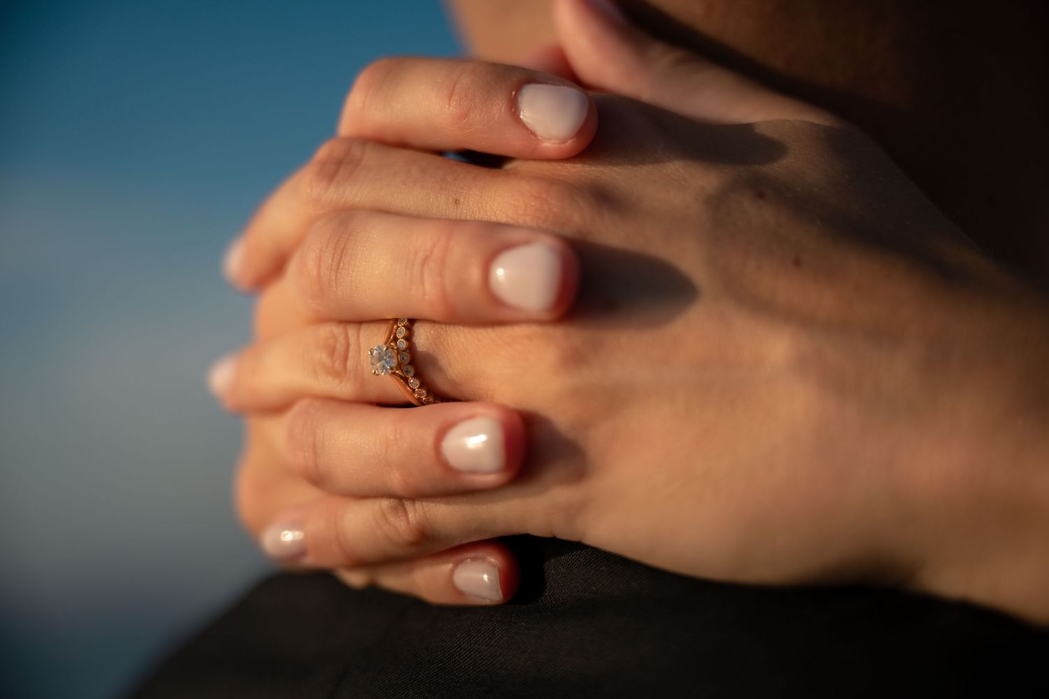 Close-up of clasped hands with a gold diamond engagement ring, bathed in warm sunlight against a blue sky.