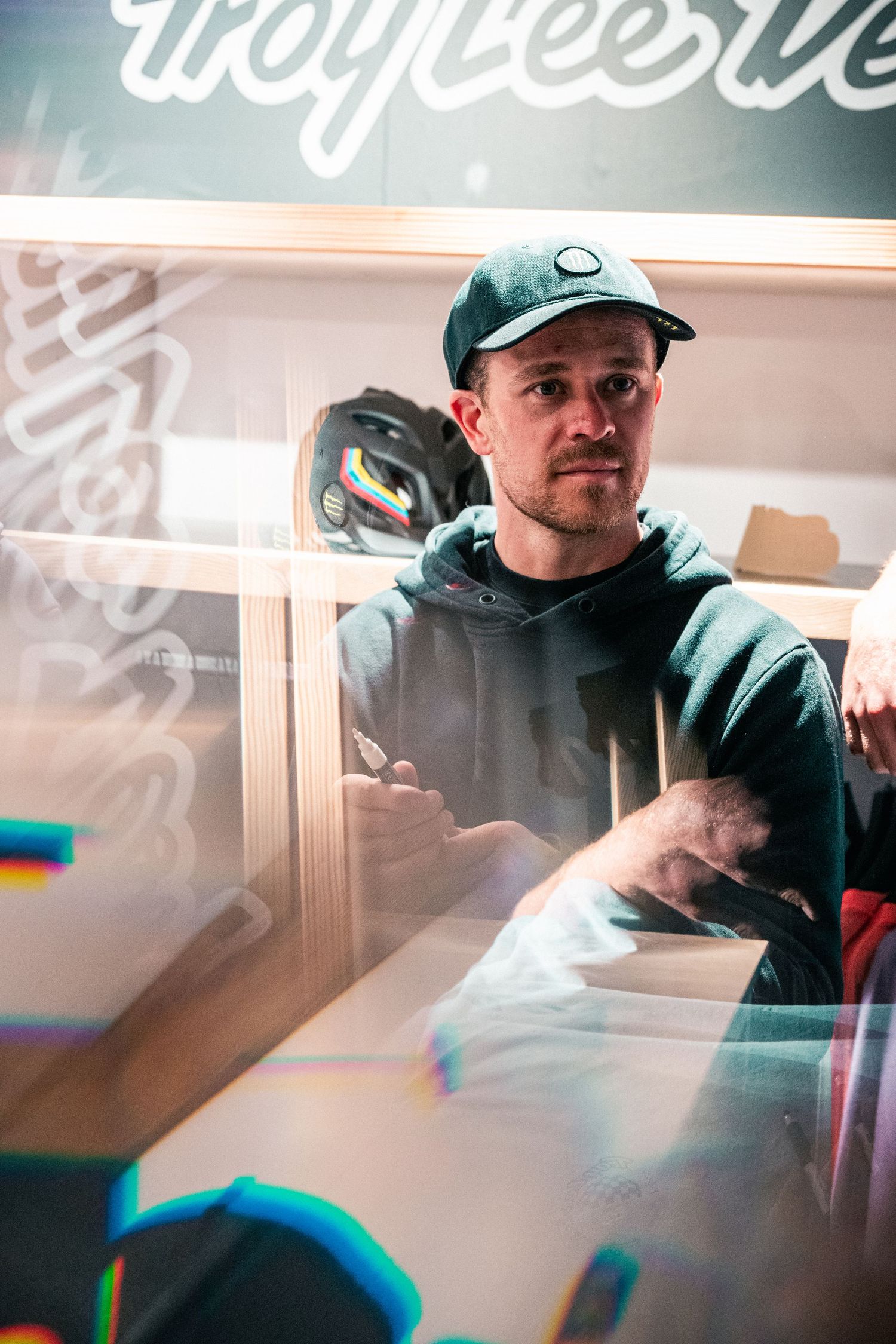 Man in green hoodie and cap stands behind glass display case in a retail store setting.