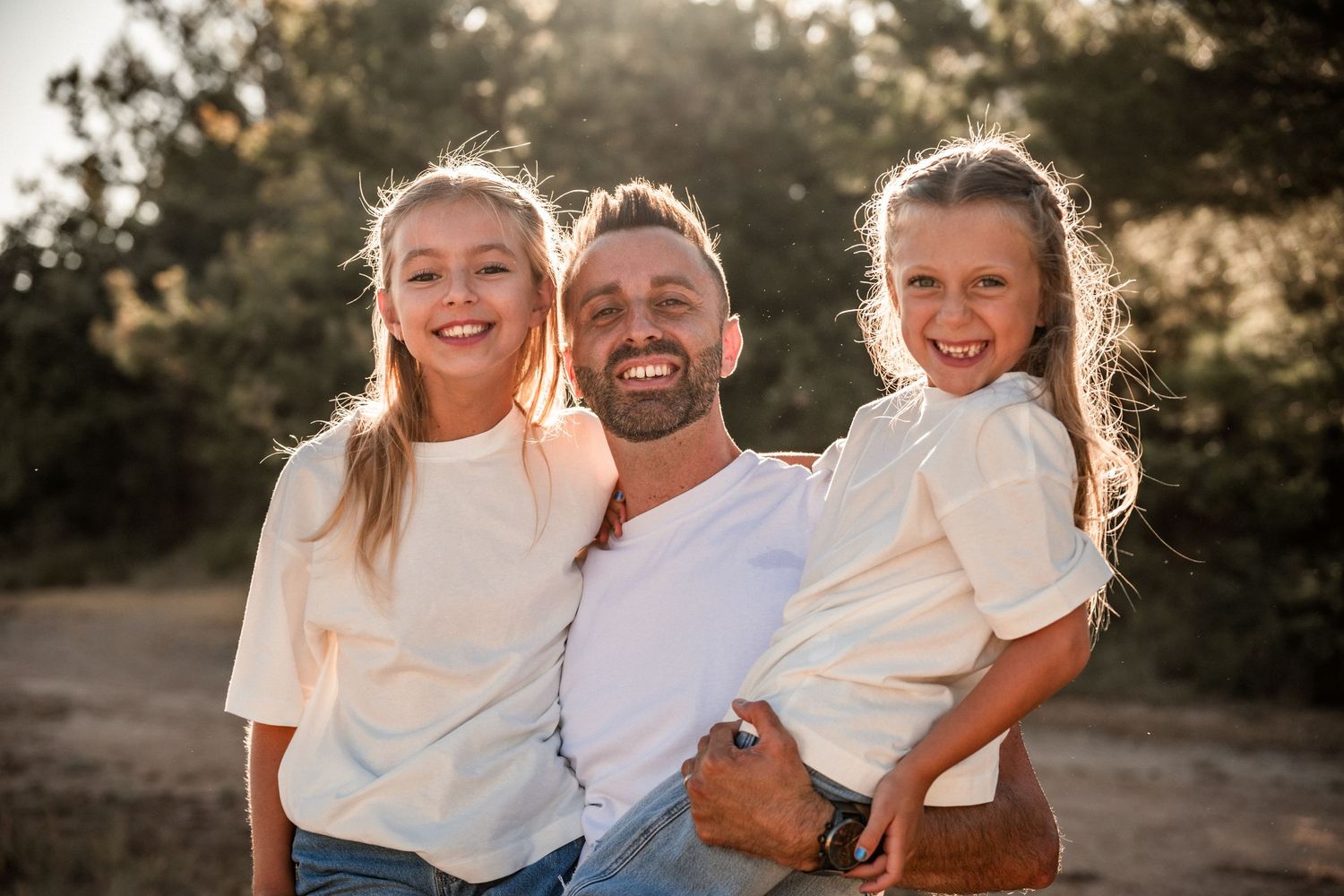 Smiling father poses outdoors with two young daughters in warm golden sunlight.