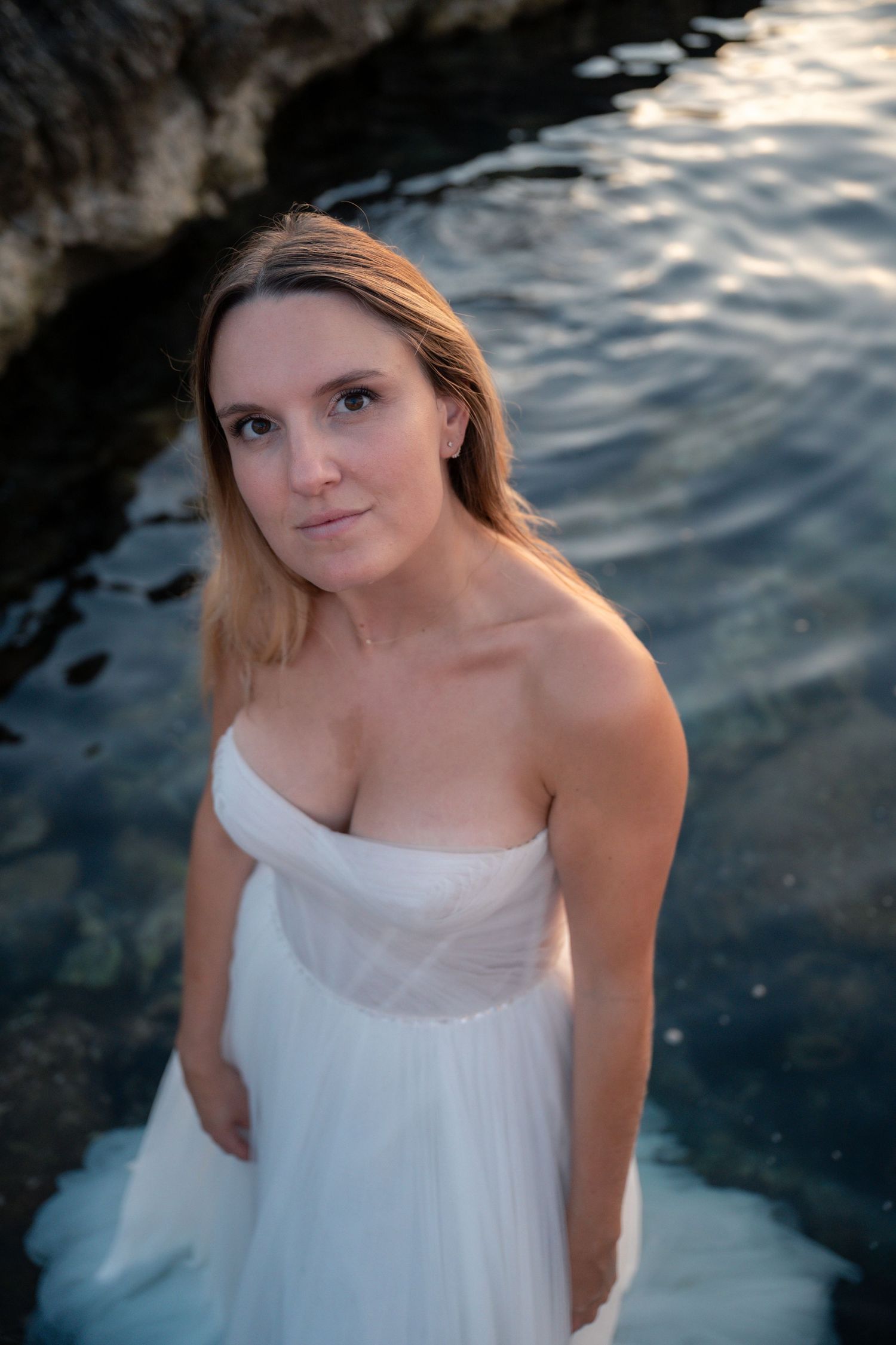 Blonde bride in strapless white gown poses by rocky ocean waters at dusk.