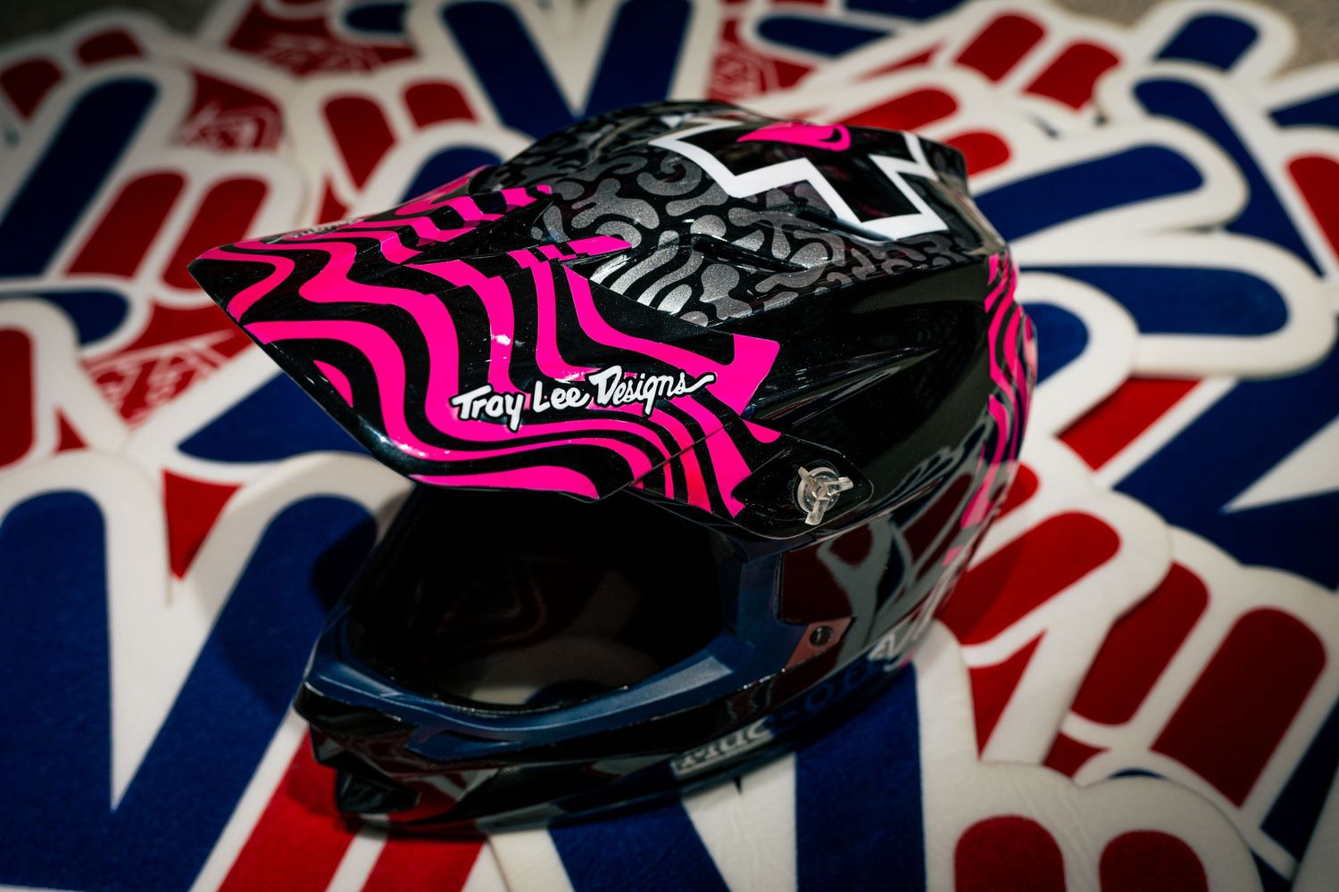 Troy Lee Designs black and pink motocross helmet displayed on colorful sticker background.