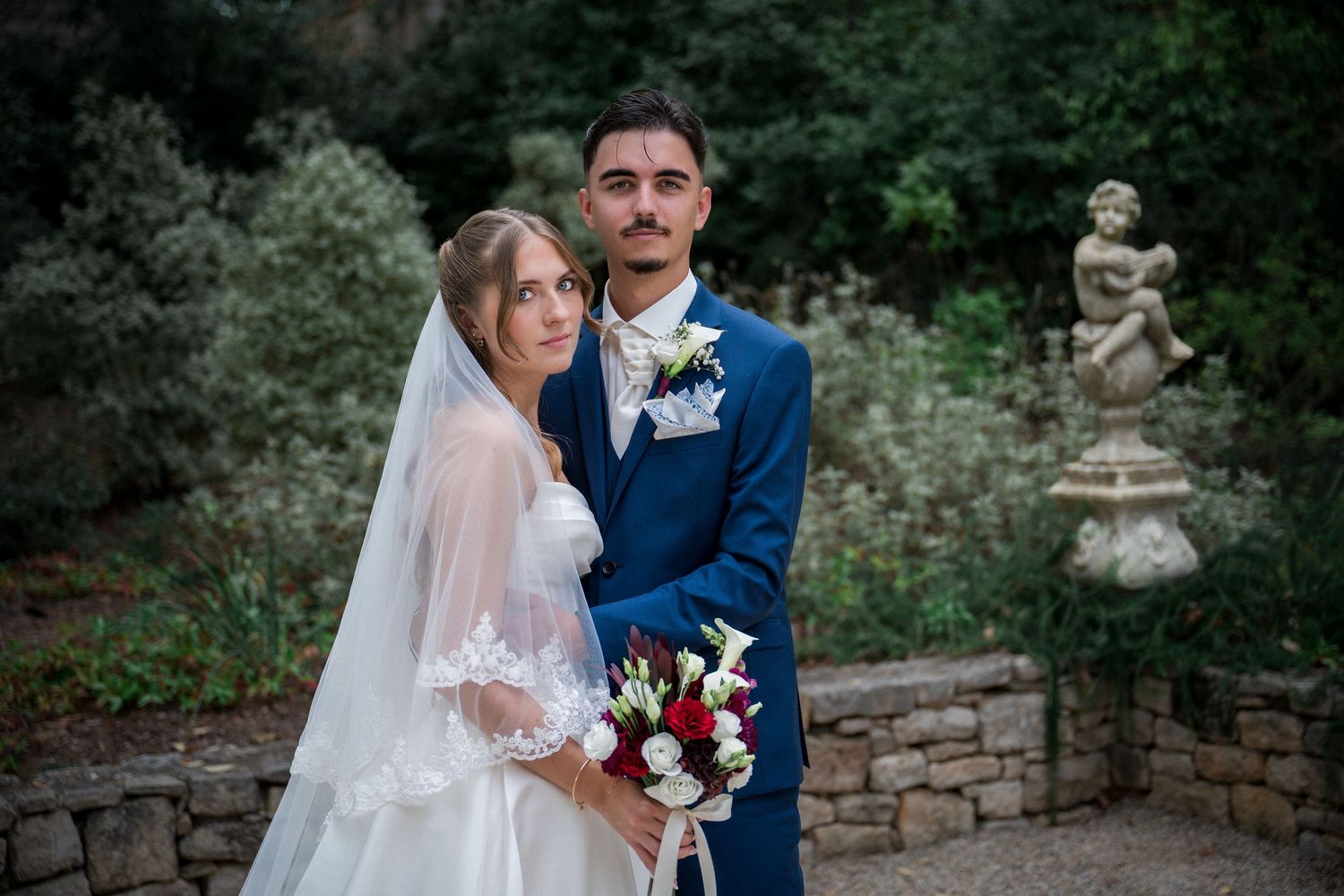 Bride and groom pose together in a garden, she wears a white veil and holds a red and white bouquet.