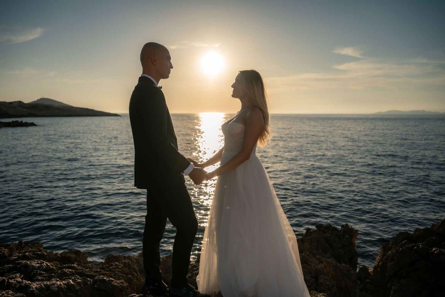 Bride and groom hold hands at sunset by the sea, silhouetted against golden light on their wedding day.