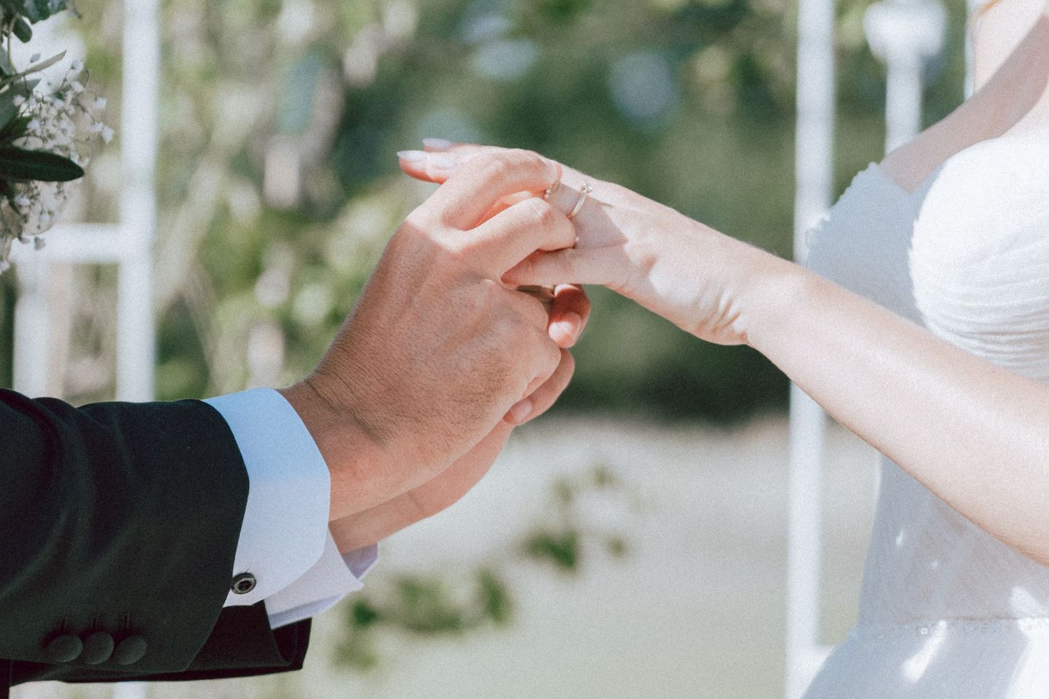 Groom placing a wedding ring on brides finger during outdoor ceremony with white floral arch backdrop.