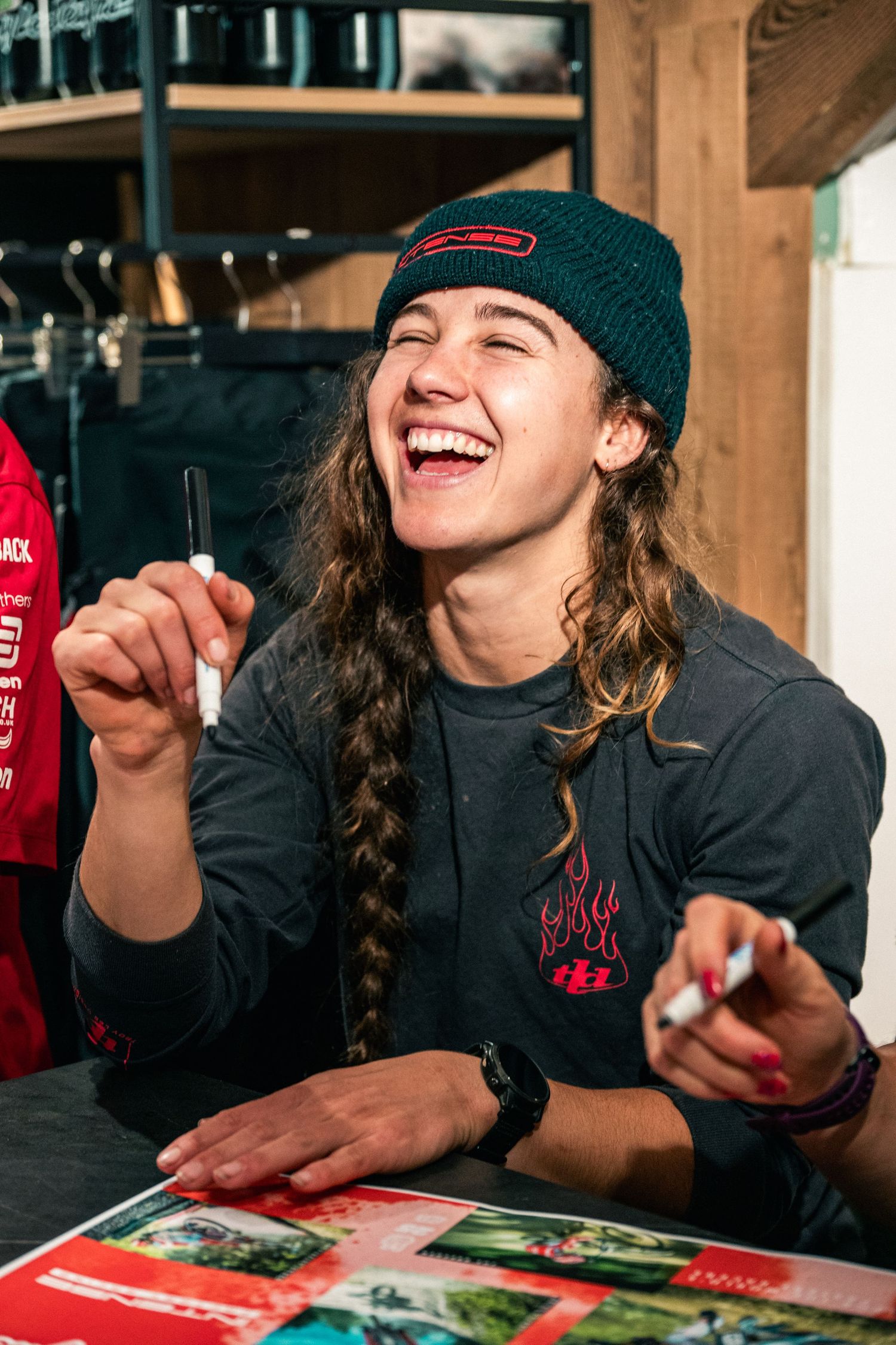 A laughing young woman in a teal beanie holds markers at a signing table indoors.