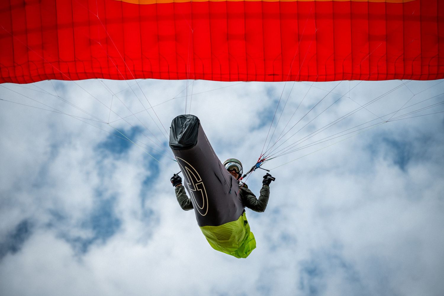 Paraglider soaring through cloudy sky with red canopy above, wearing black jacket and yellow pants.