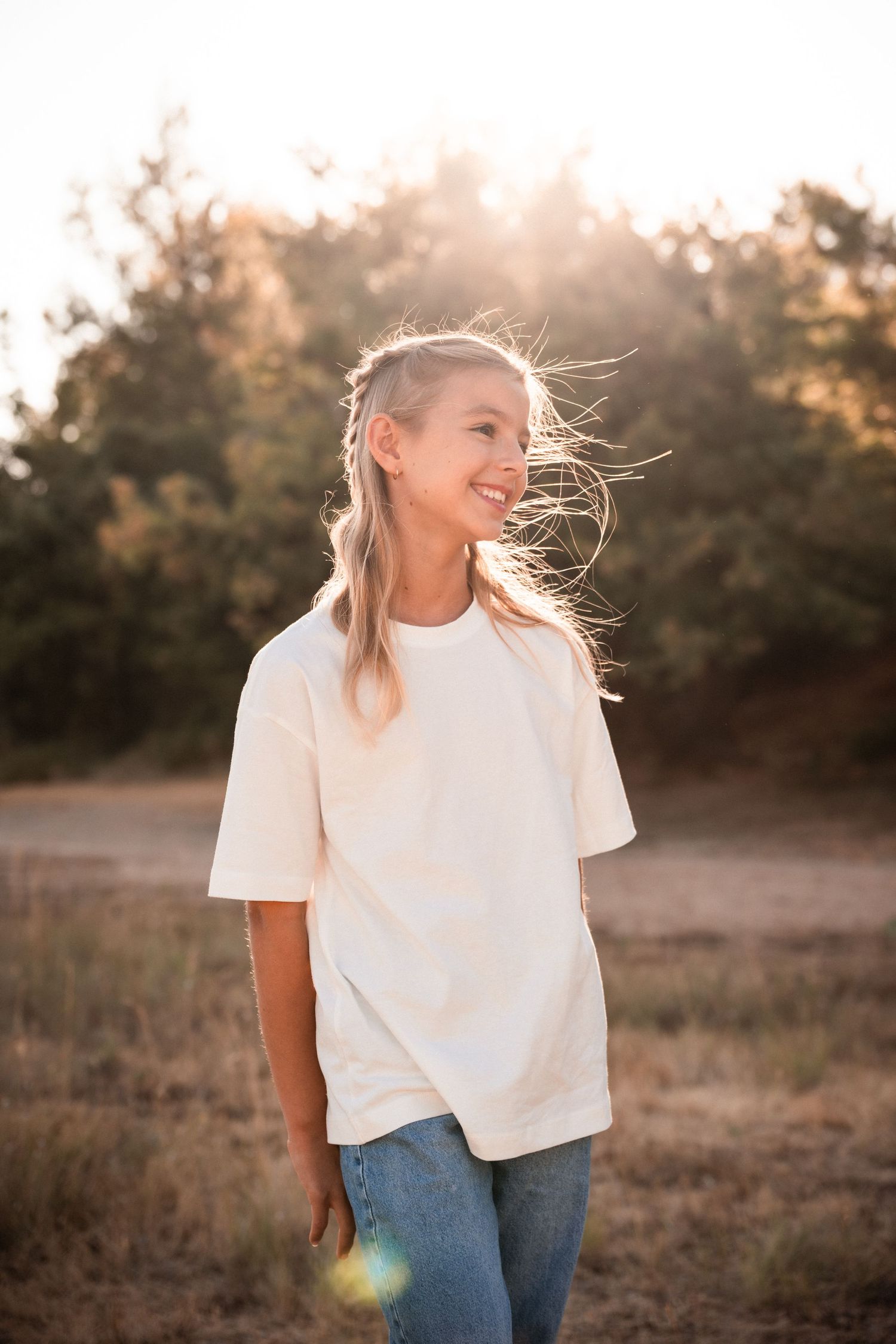 Cheerful blonde girl in white oversized tee standing in a sun-drenched field.
