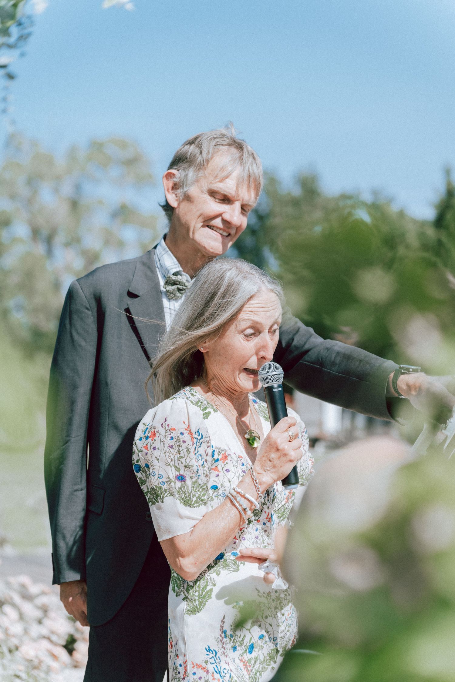 Older couple sharing a joyful moment with microphone at sunny outdoor wedding celebration.