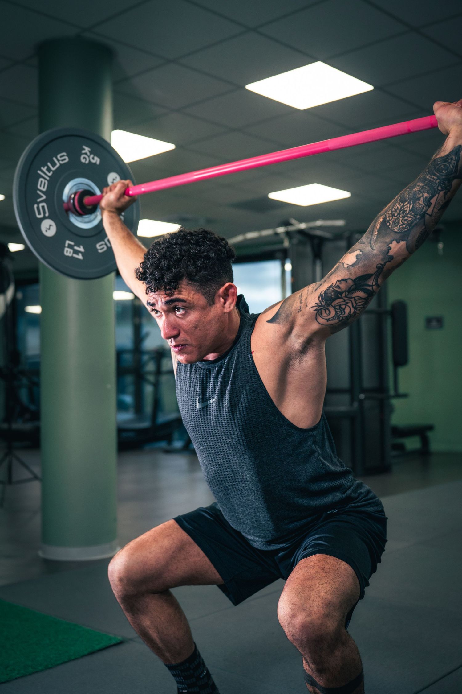 Tattooed muscular man performing overhead barbell squat exercise in modern gym.