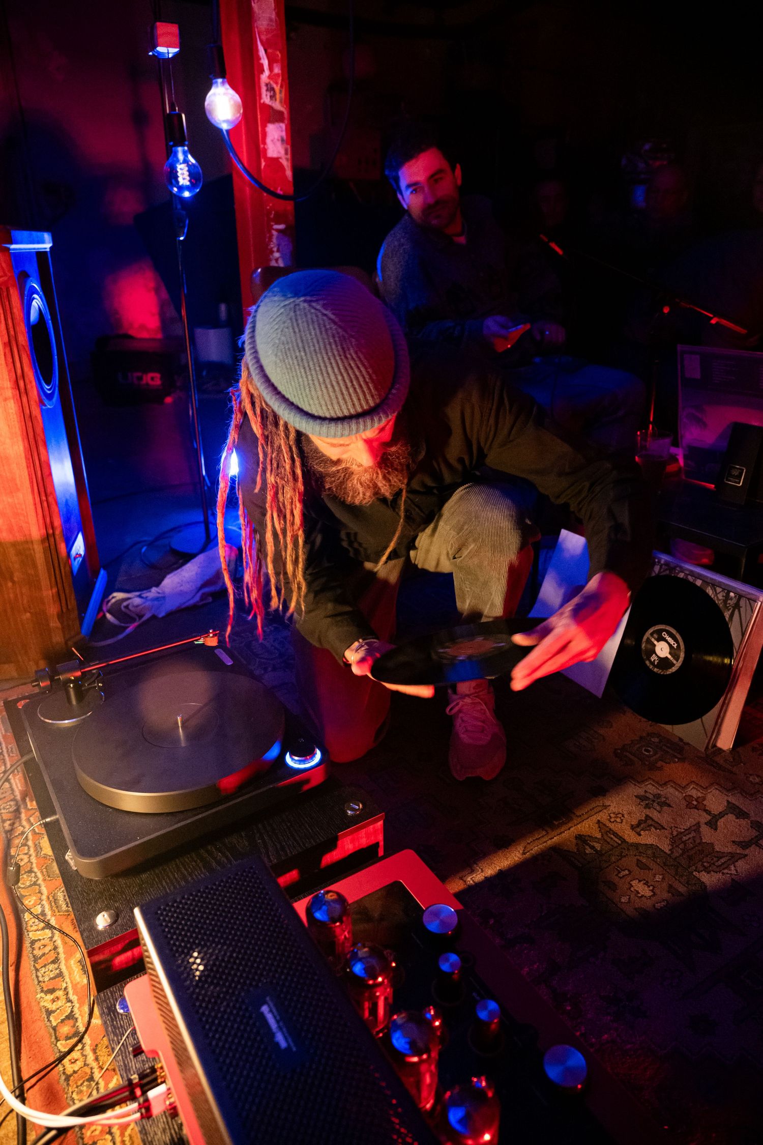 DJ with dreadlocks and beanie hat mixing vinyl records on turntables in a dimly lit red and blue neon venue.