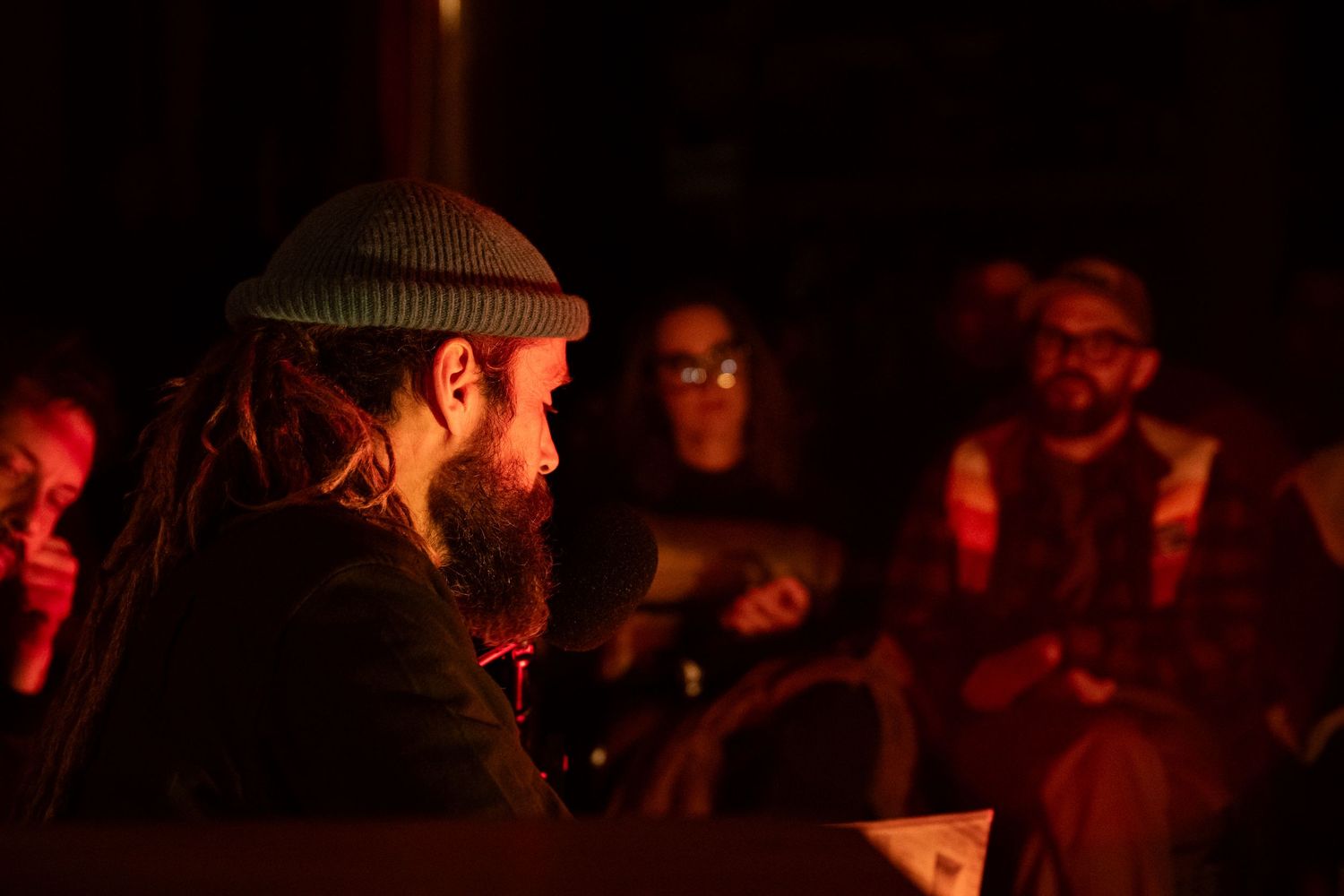 Bearded man with dreadlocks and beanie hat in dimly lit gathering with people in background.