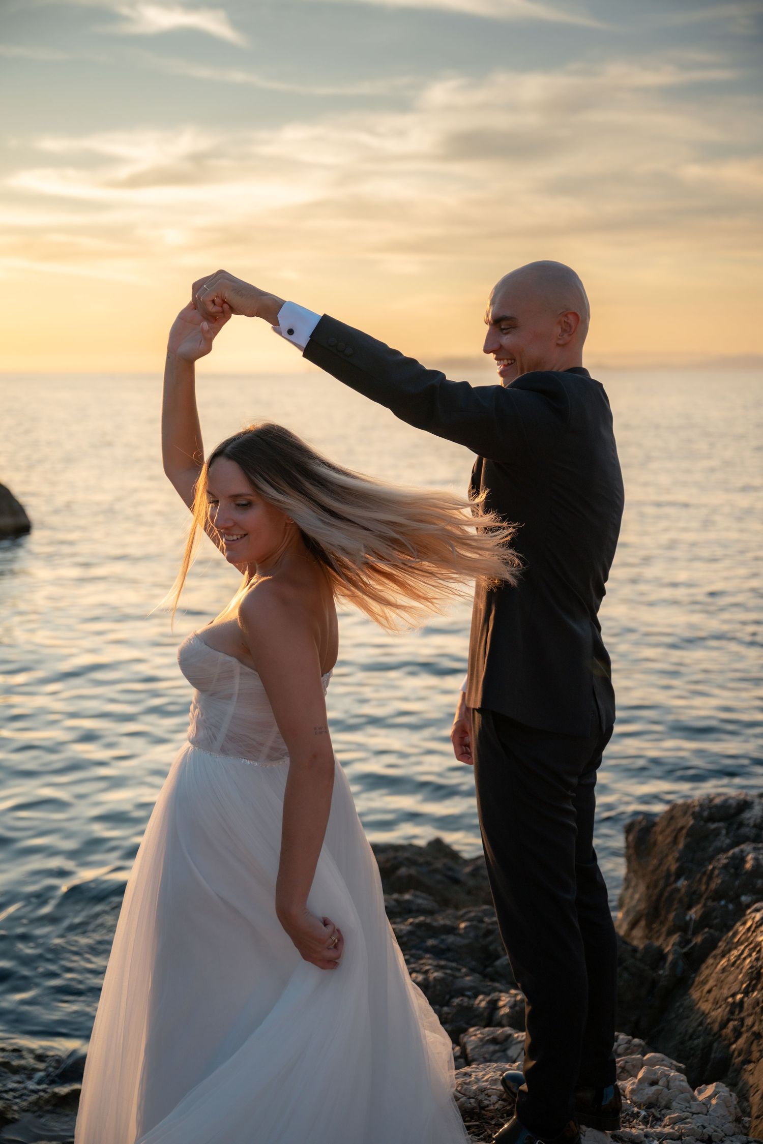 Groom twirling bride in white dress on rocky seaside during stunning sunset.