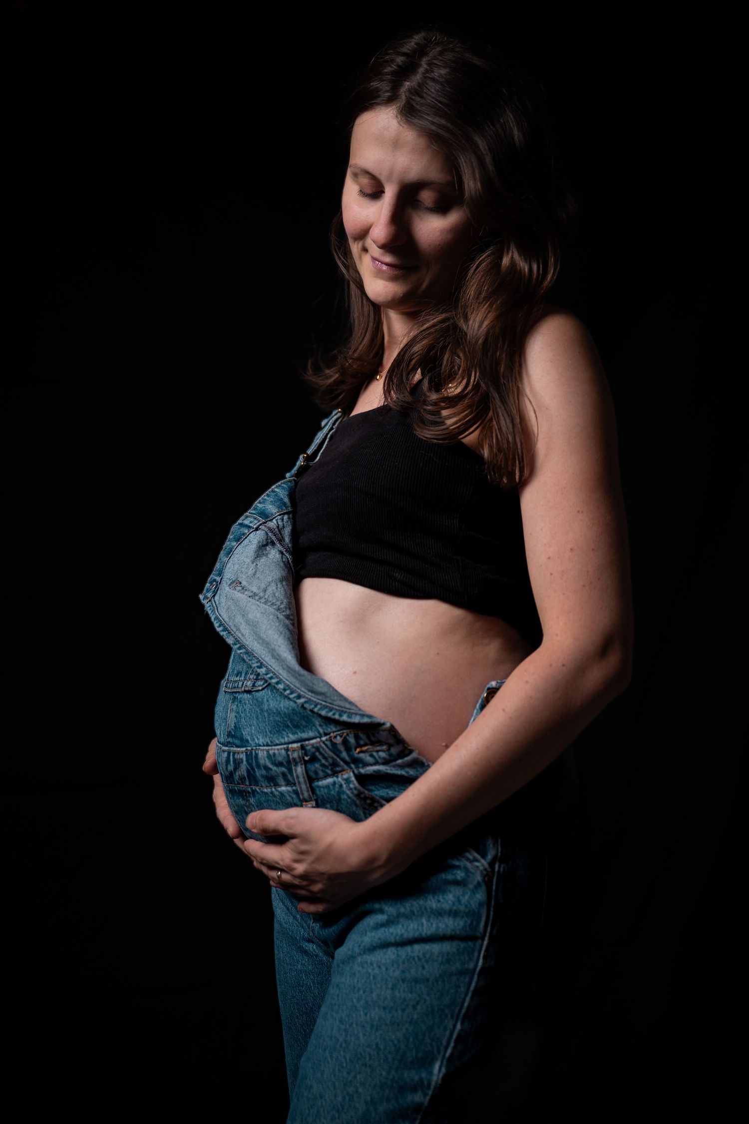 Smiling pregnant woman in denim overalls and black crop top cradling her baby bump against a dark background.
