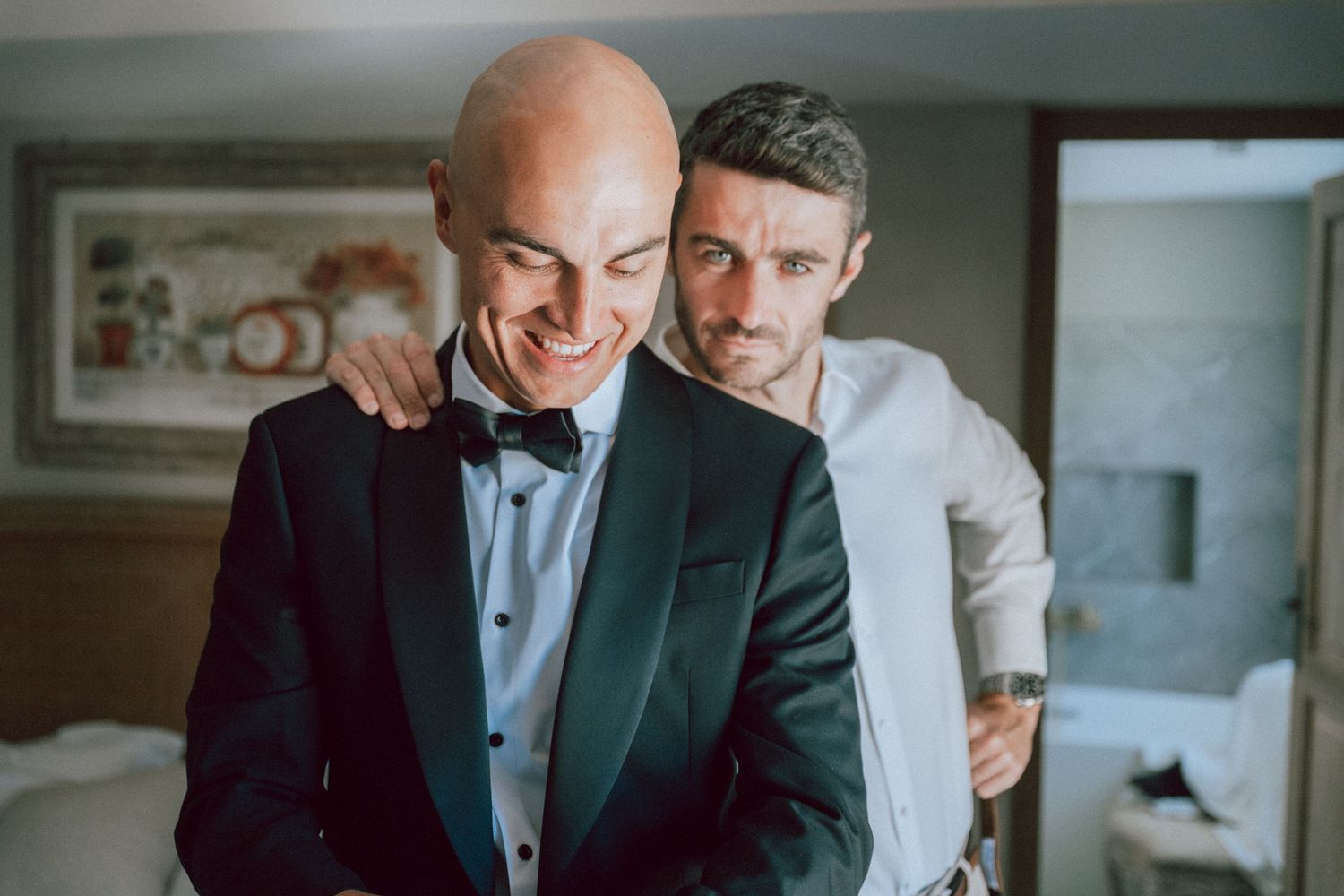 Groomsman helping bald groom in dark tuxedo with bow tie prepare for wedding ceremony.