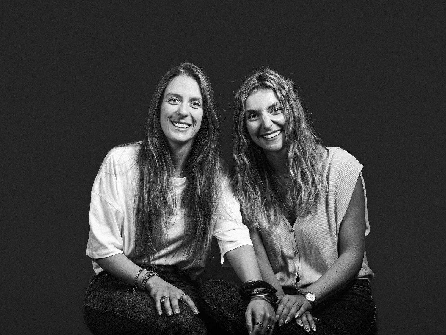 Two smiling women with long hair sit together in a black and white portrait photo against a dark background.