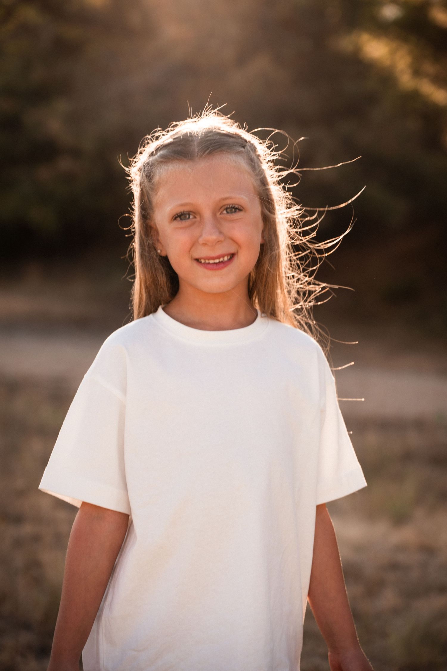Smiling young girl with windswept hair wearing an oversized white t-shirt outdoors in golden sunlight.