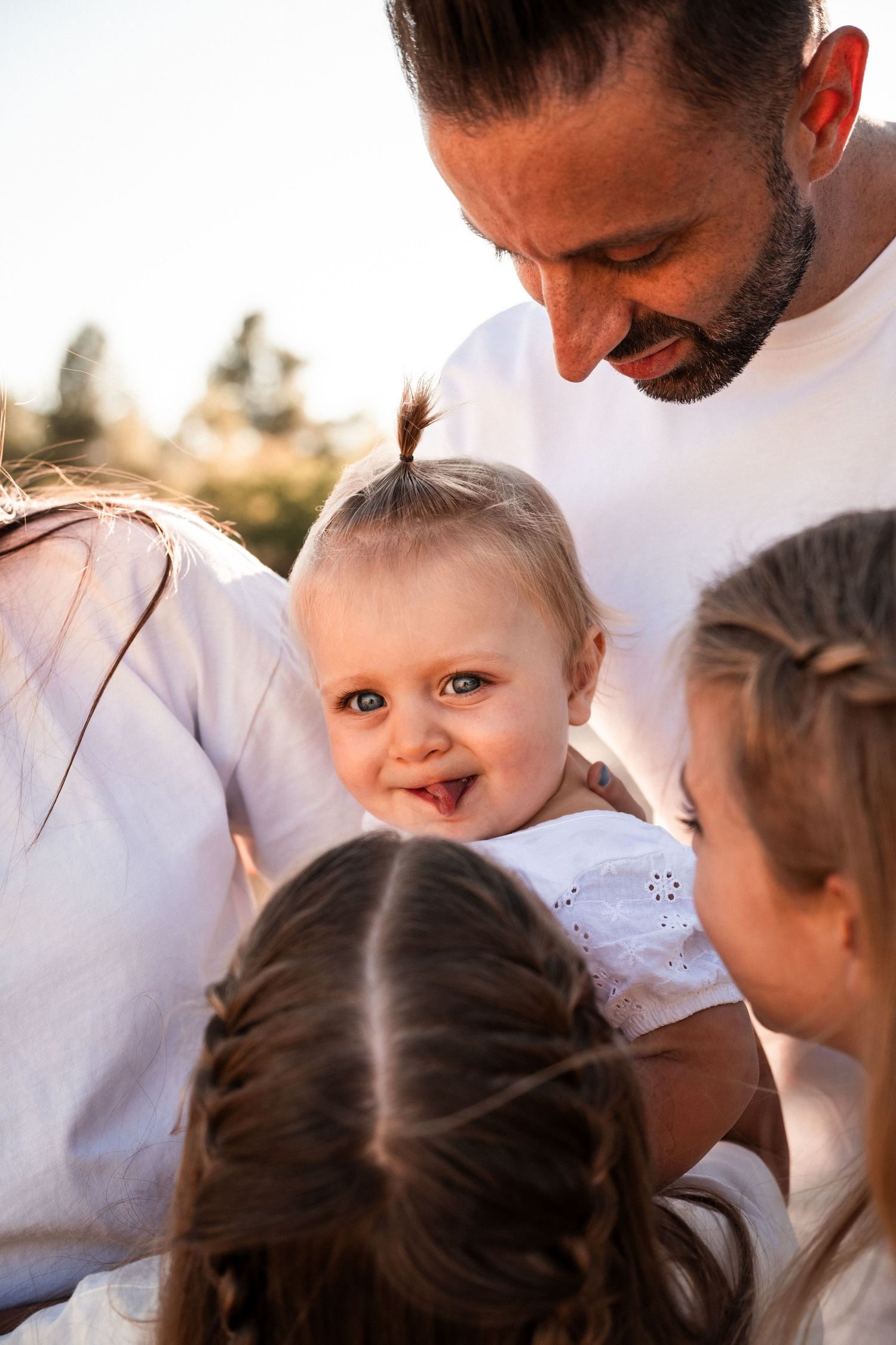 A smiling baby with bright green eyes is held by a bearded man in white, surrounded by two young girls outdoors.