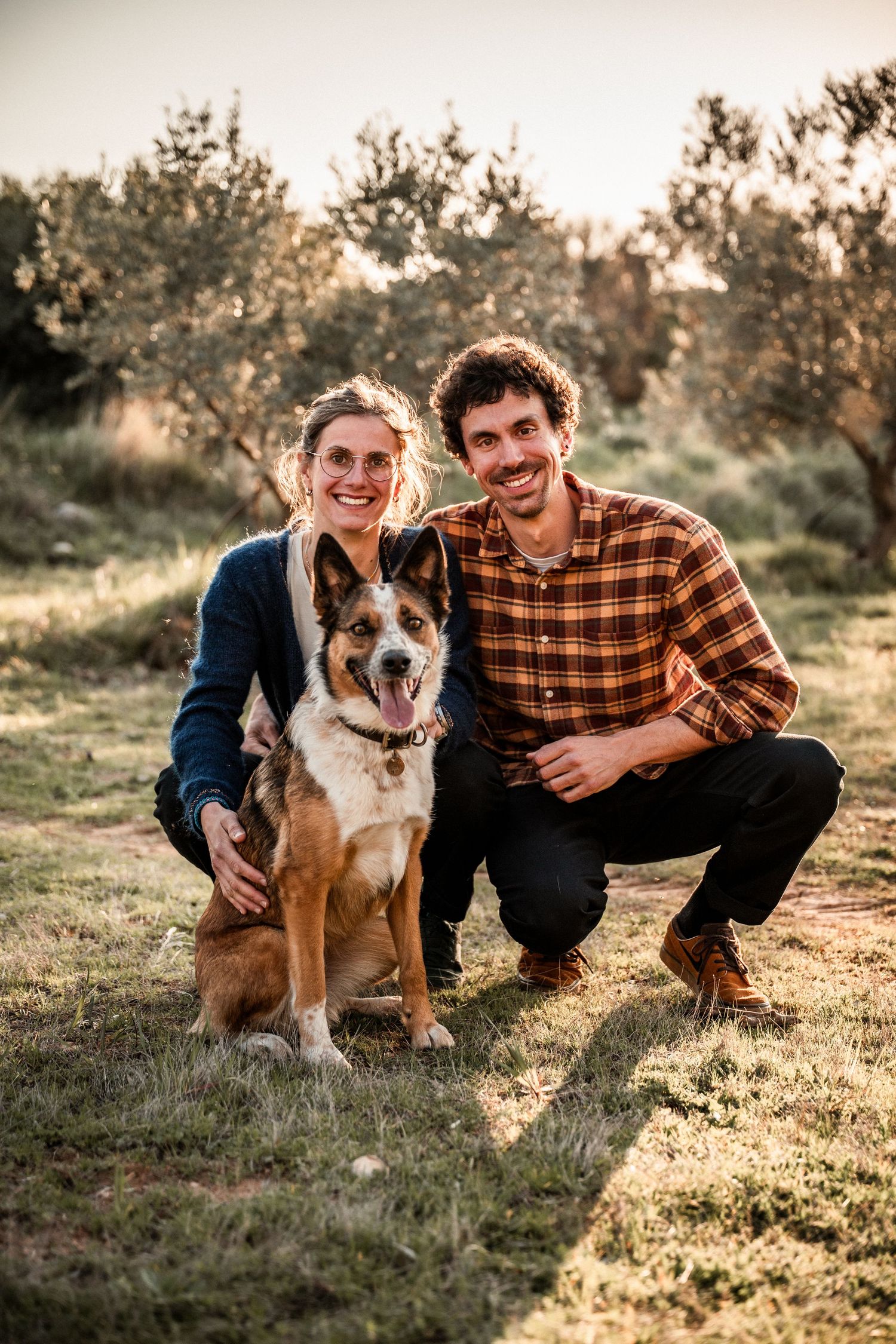Smiling couple crouching with their happy German Shepherd dog in a sunny olive grove.