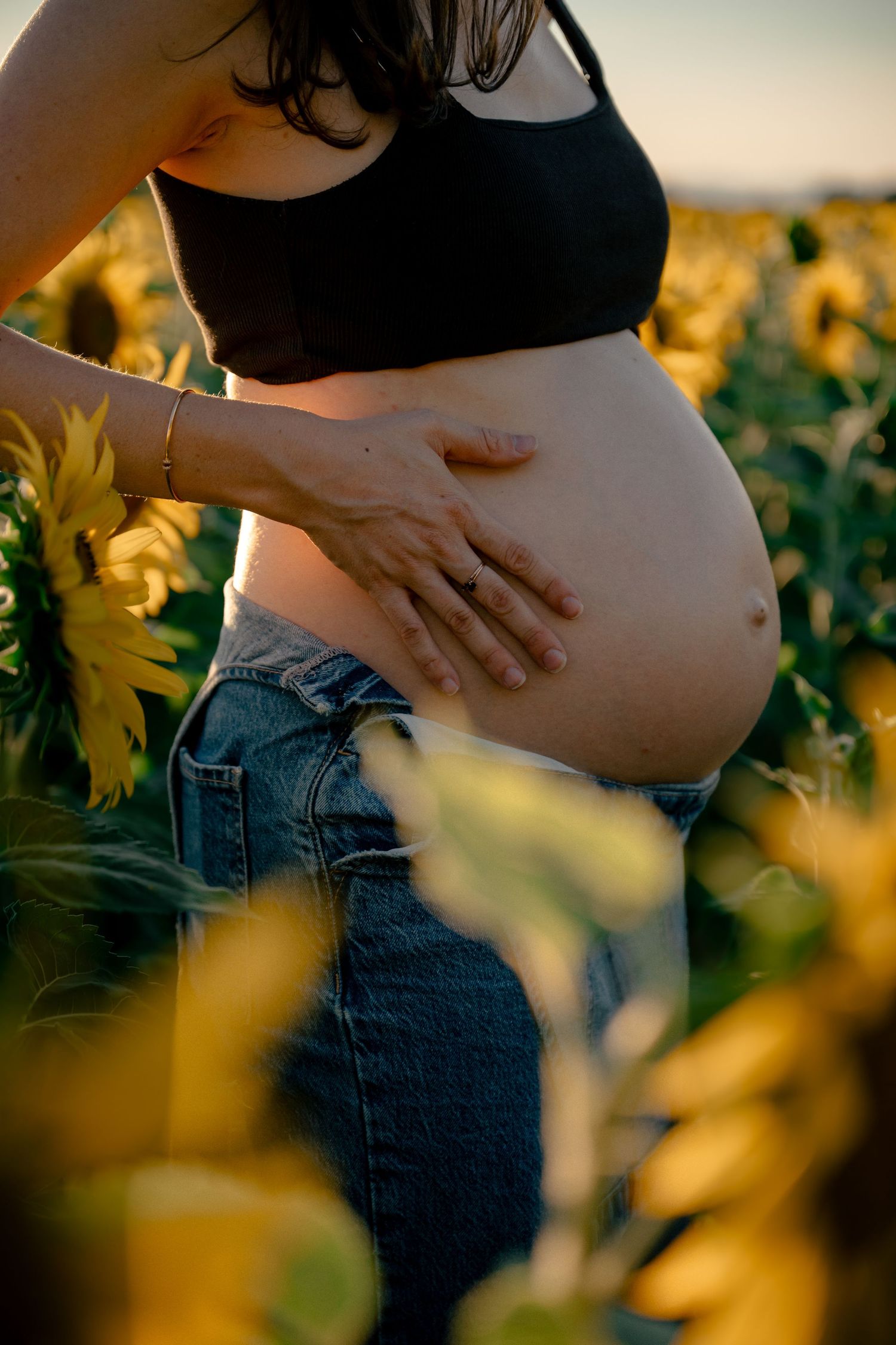 Close-up of pregnant woman in black crop top holding her bare baby bump surrounded by blooming sunflowers at sunset.