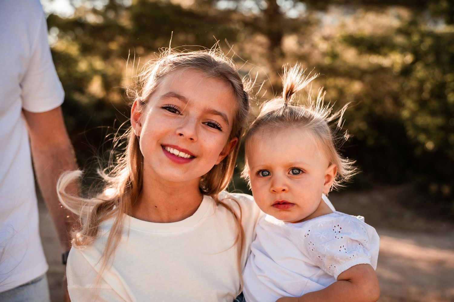 Older sister smiles holding baby sibling in warm golden hour outdoor portrait.