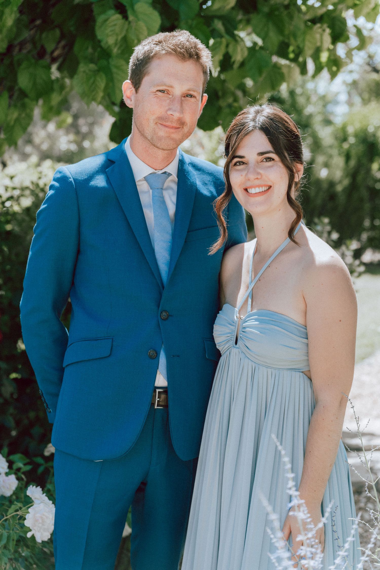 Groomsman in blue suit poses with bridesmaid in light blue gown outdoors at wedding.