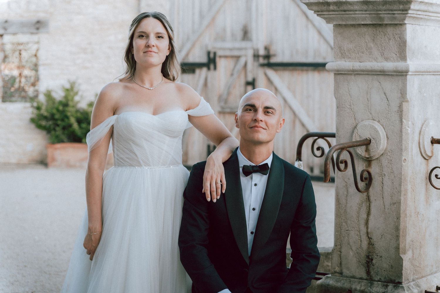 Bride standing behind seated groom near stone fountain, both in formal wedding attire.