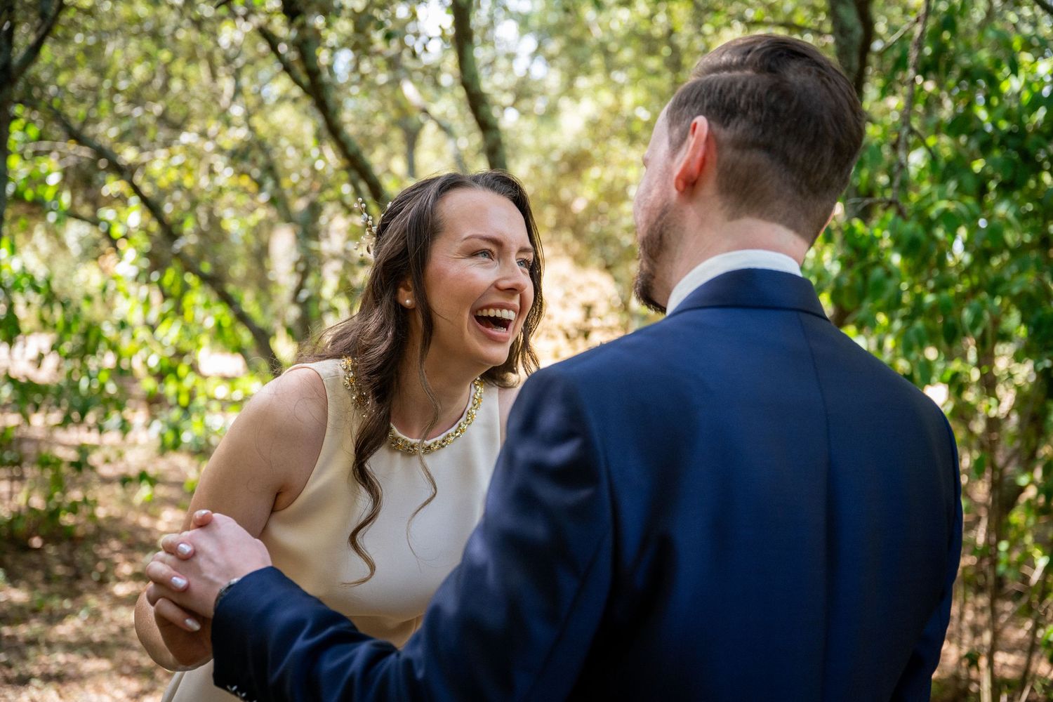 Bride laughing joyfully with groom in navy suit during outdoor wedding ceremony surrounded by lush green trees.